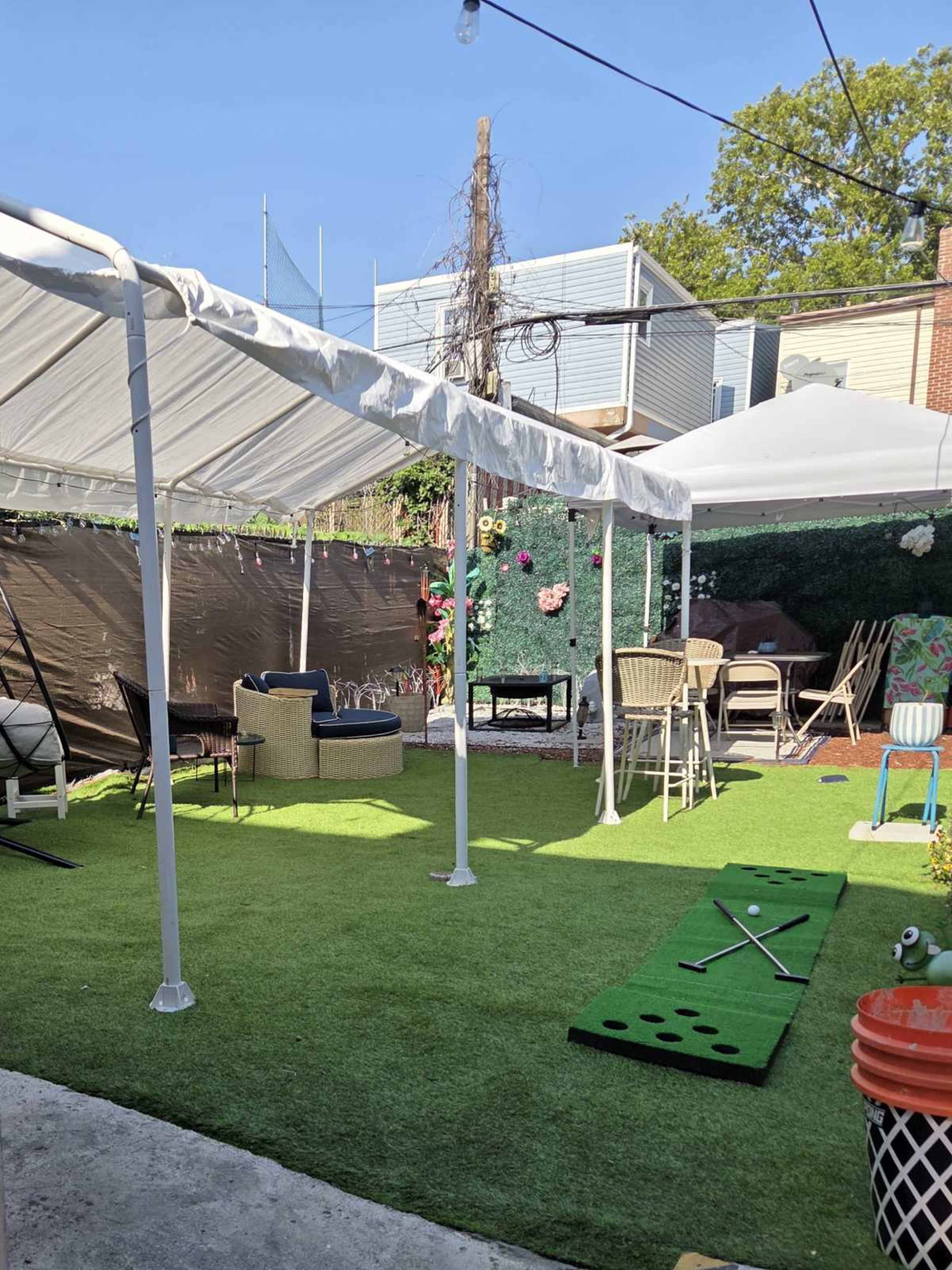 The scene shows a backyard area with artificial grass, tents, chairs, and a mini-golf setup, surrounded by greenery and a fence.