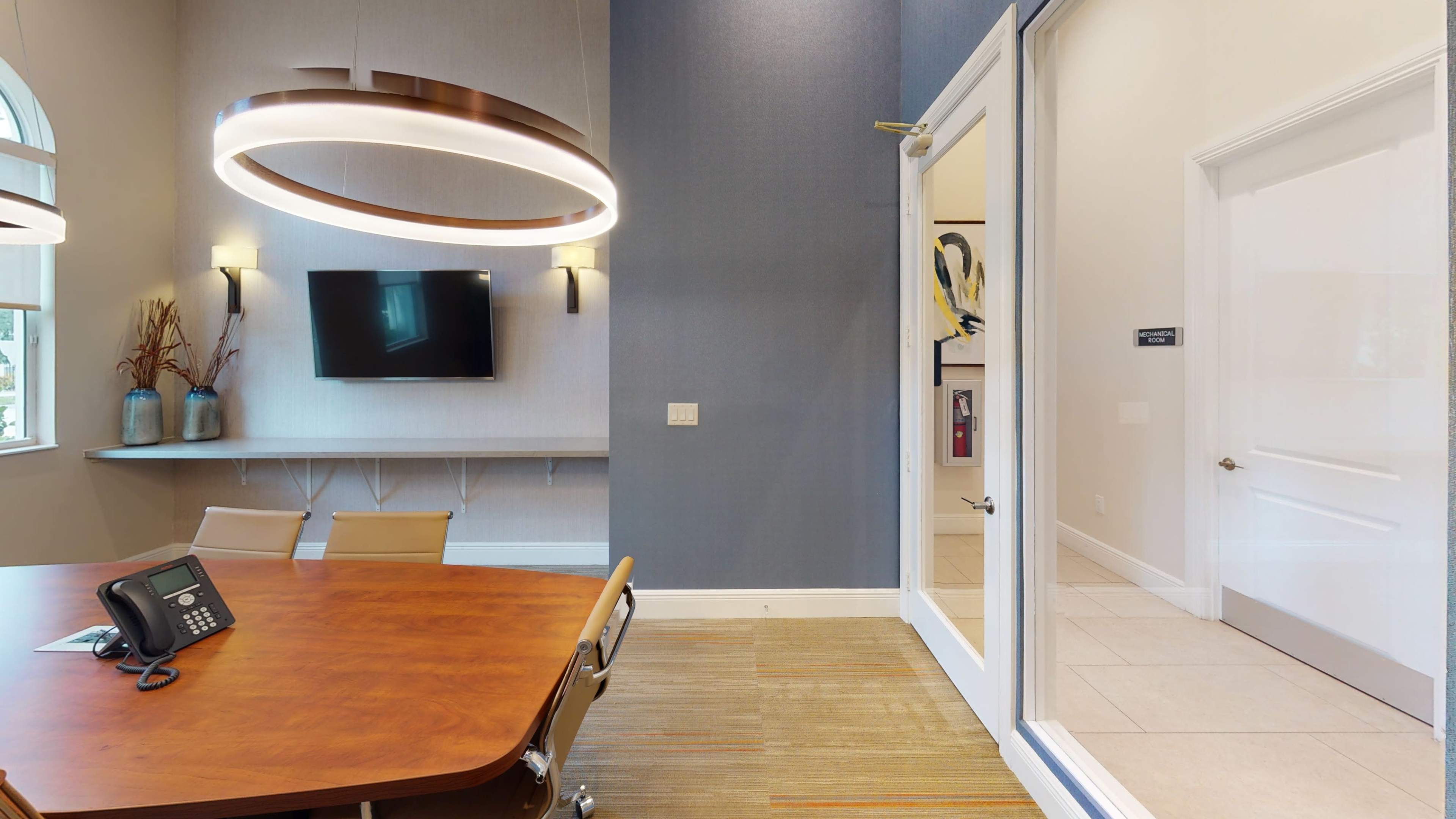 A modern conference room with a wooden table, ergonomic chairs, a mounted television, and a glass door leading to another room.