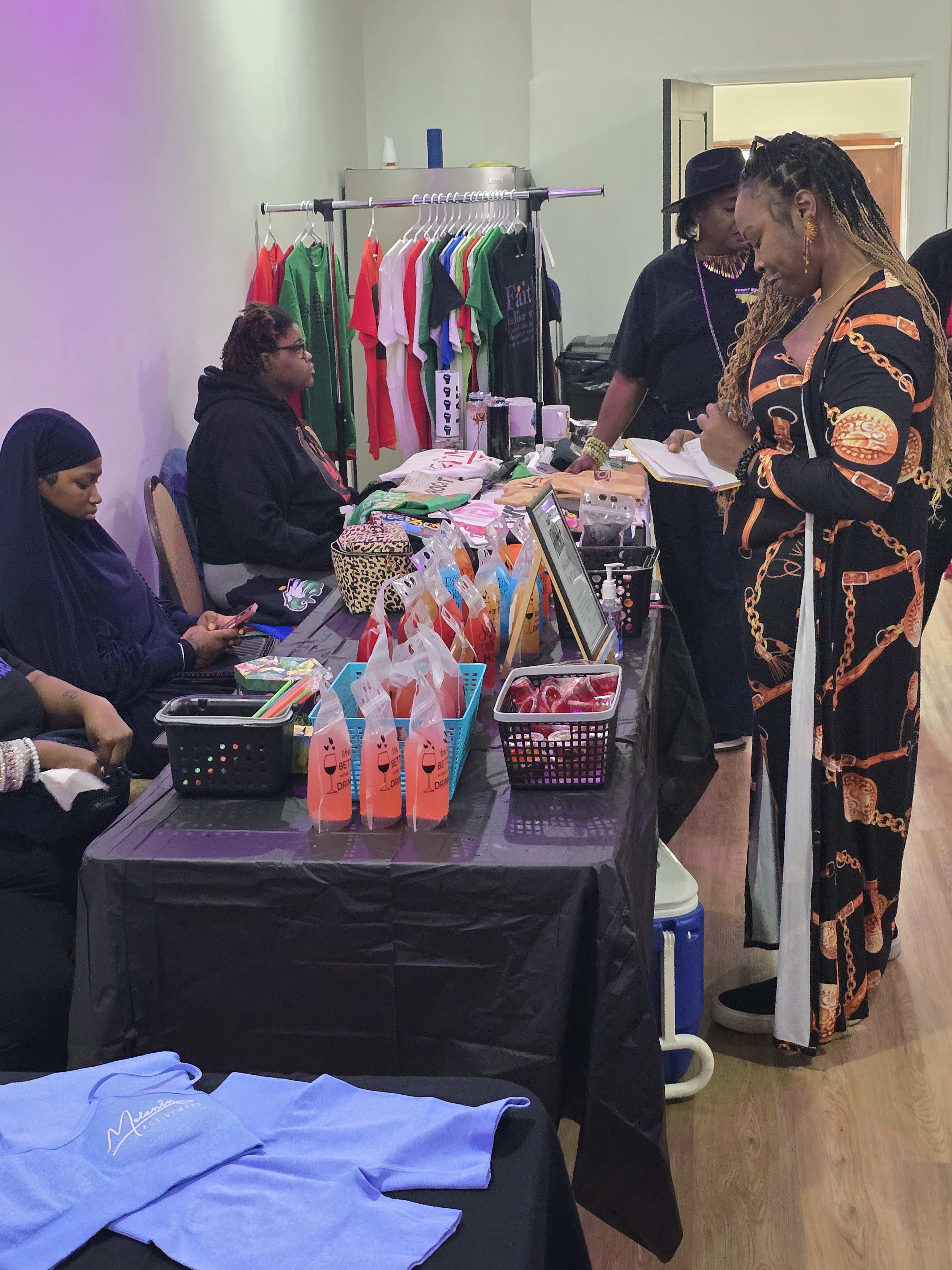 A diverse group of people is gathered at a vendor table showcasing clothing and accessories in a well-lit indoor space.