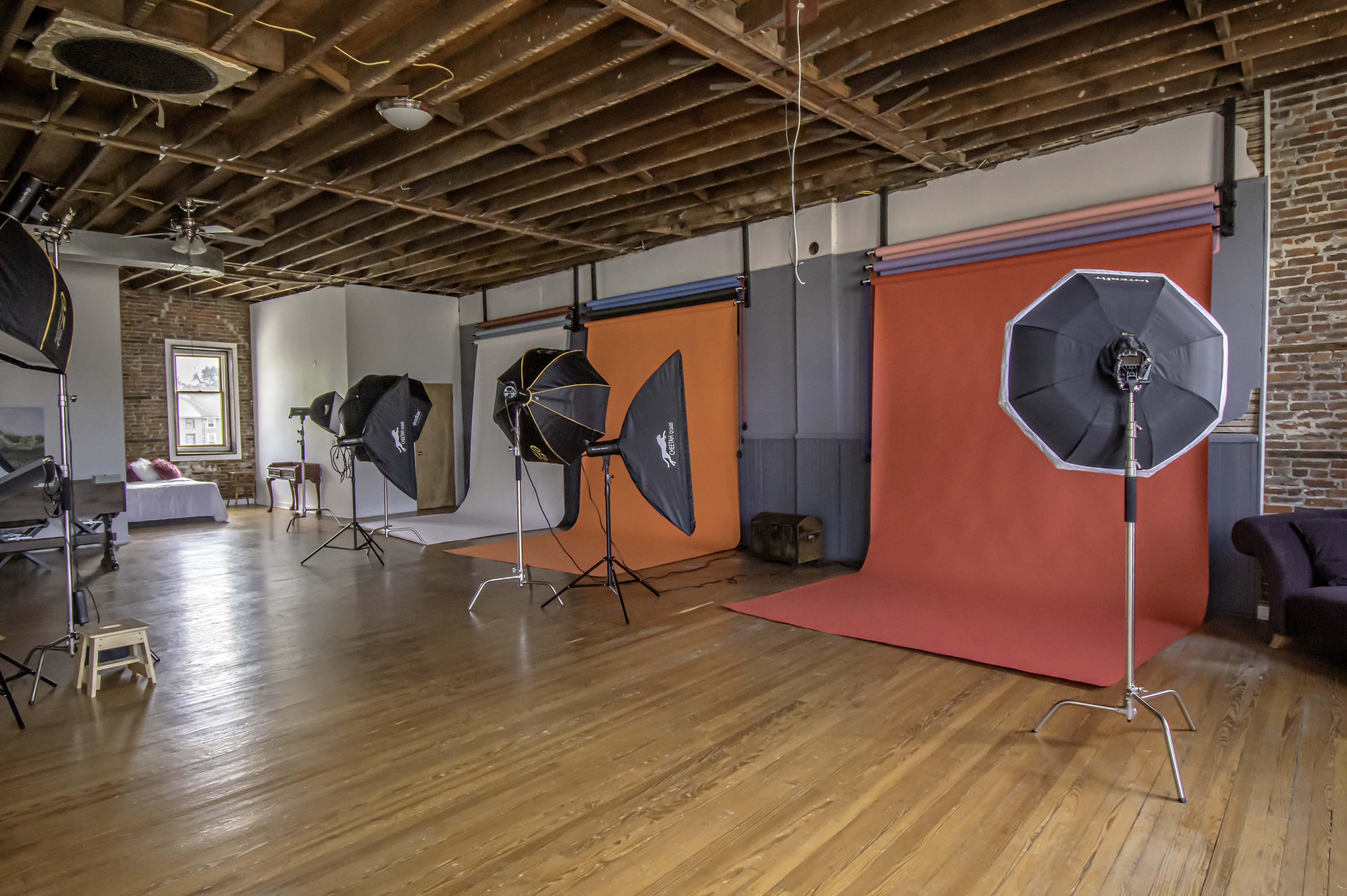 A photography studio with multiple backdrops and lighting equipment set up in a spacious room featuring exposed wooden floors and a high ceiling.