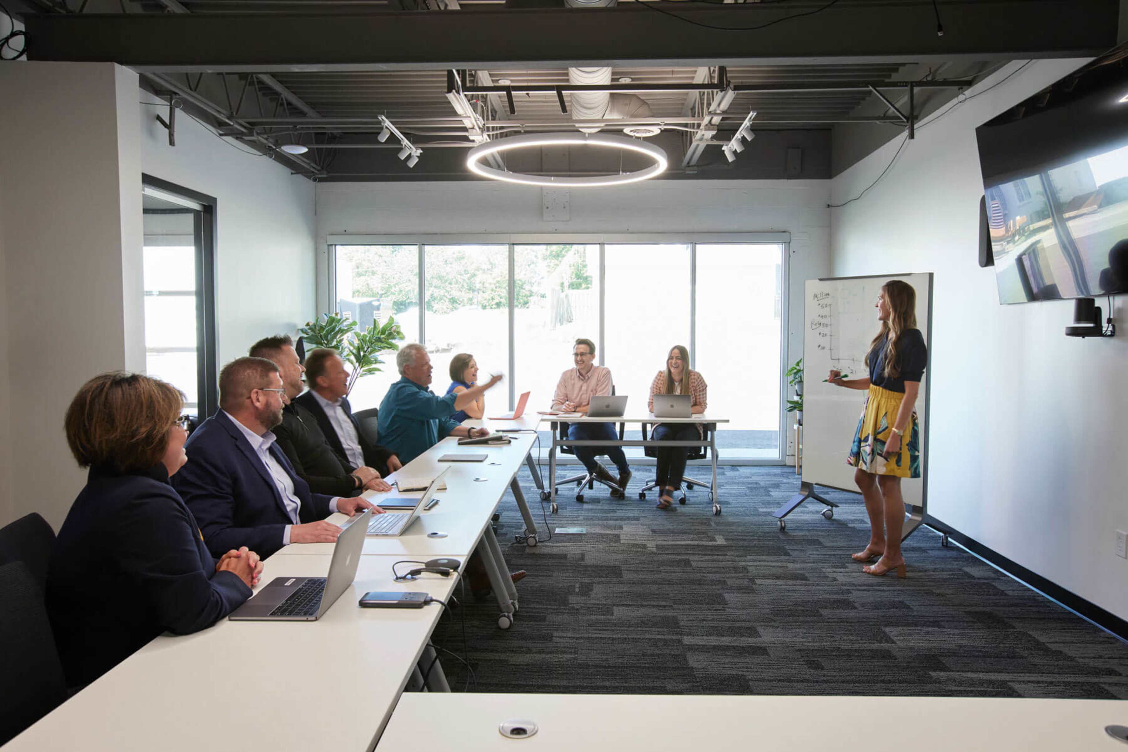 A group of eight professionals is seated at a long table in a modern conference room, while a woman stands at the front presenting to the team.