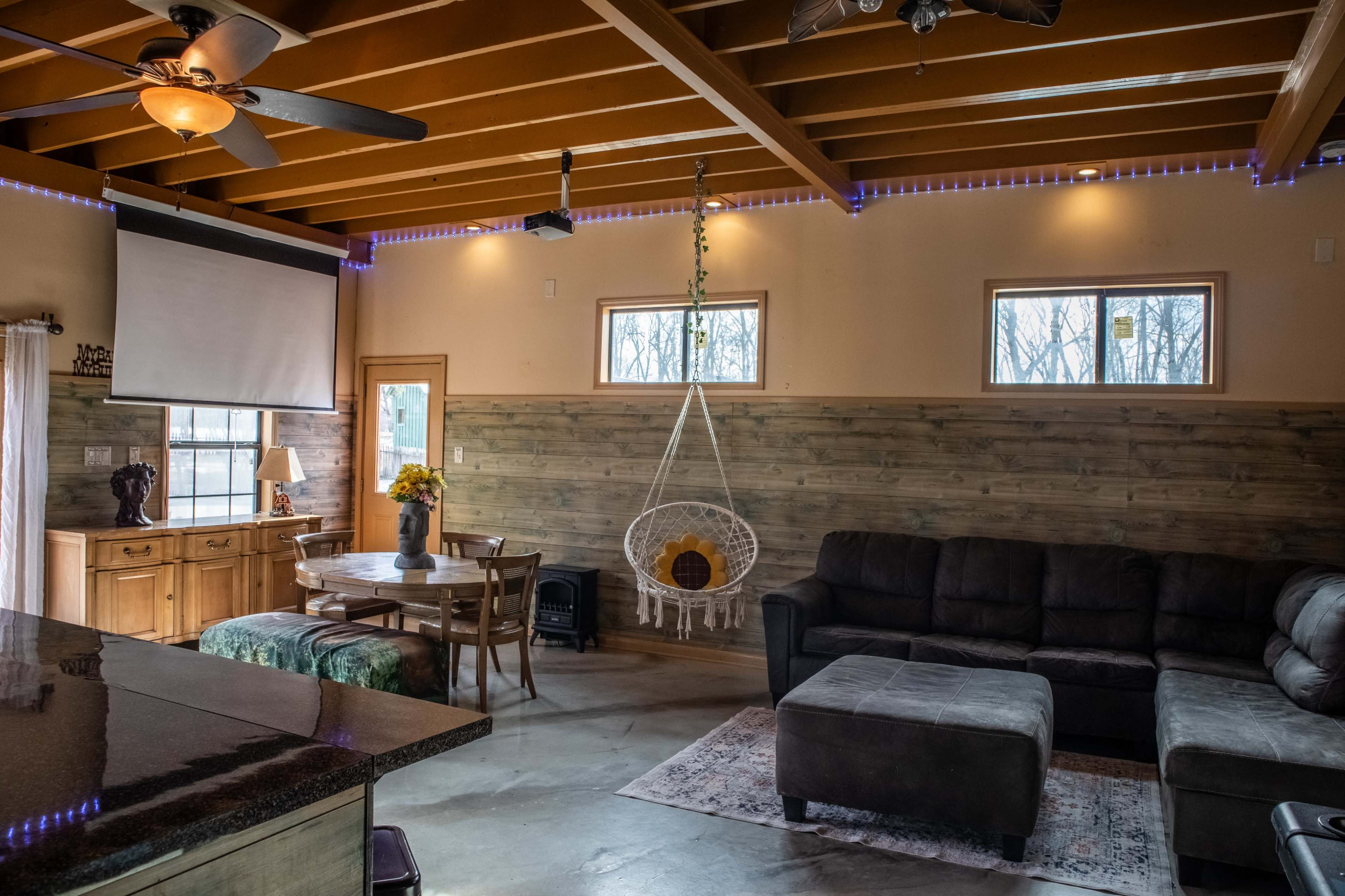 The image shows a cozy living room with a sectional sofa, a hanging chair, and a wooden dining table, all set against a backdrop of wood-paneled walls and windows.