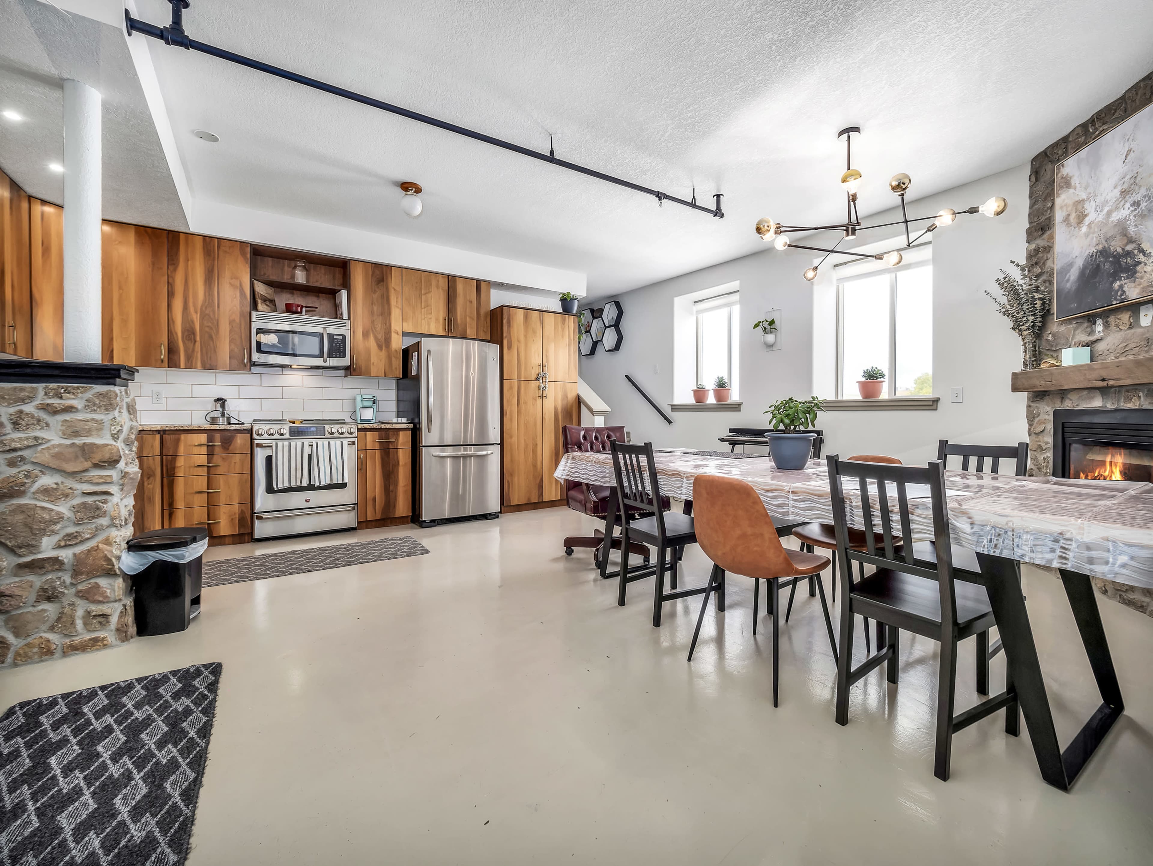 A modern kitchen and dining area featuring wooden cabinetry, a metal refrigerator, a dining table surrounded by various chairs, and a stone fireplace.