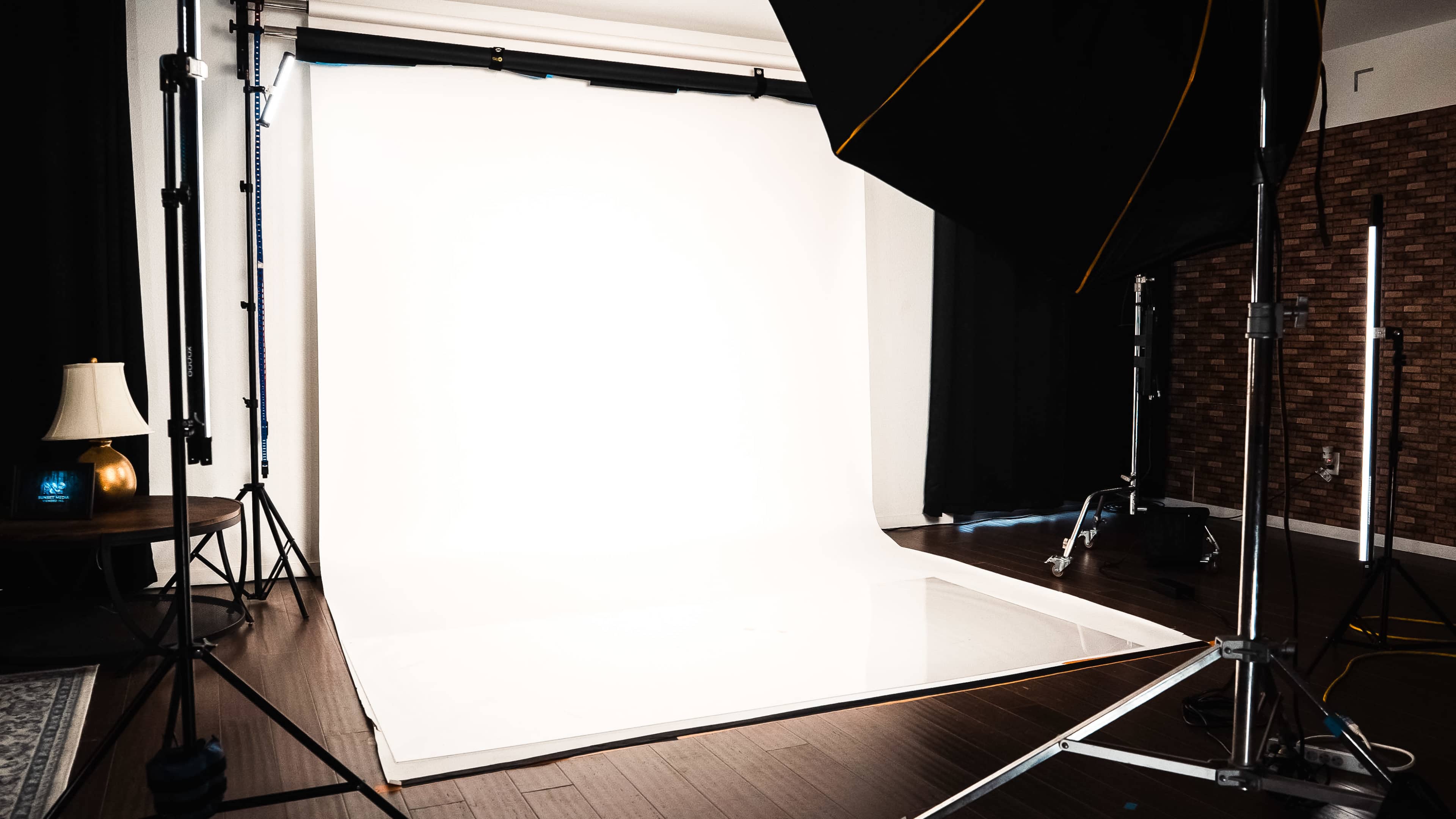 The image shows a photography studio setup with a large white backdrop, lighting equipment, and a small table with a lamp.