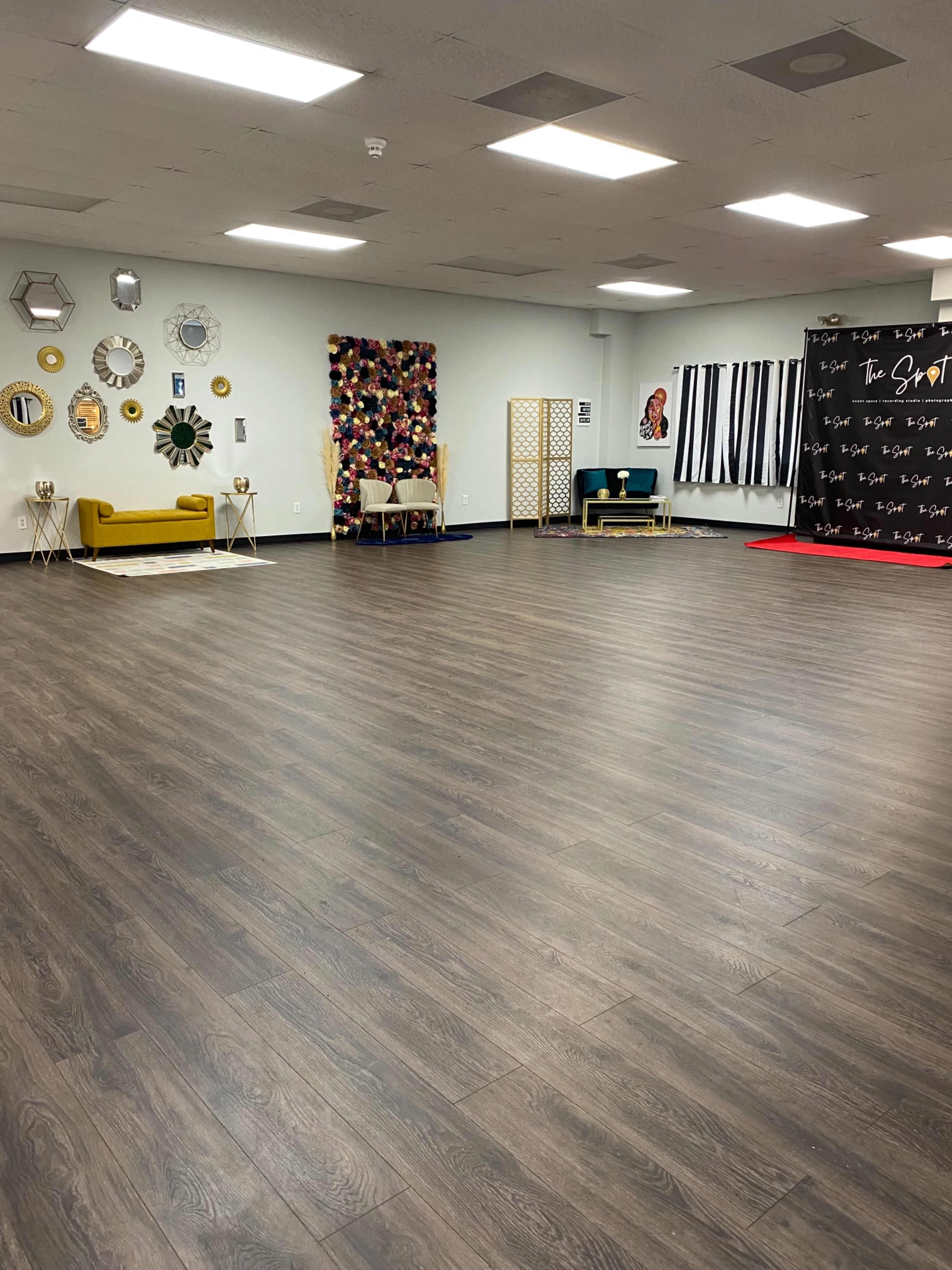 A spacious, empty room with wooden flooring, decorated walls featuring various wall hangings, seating areas, and a backdrop for events.