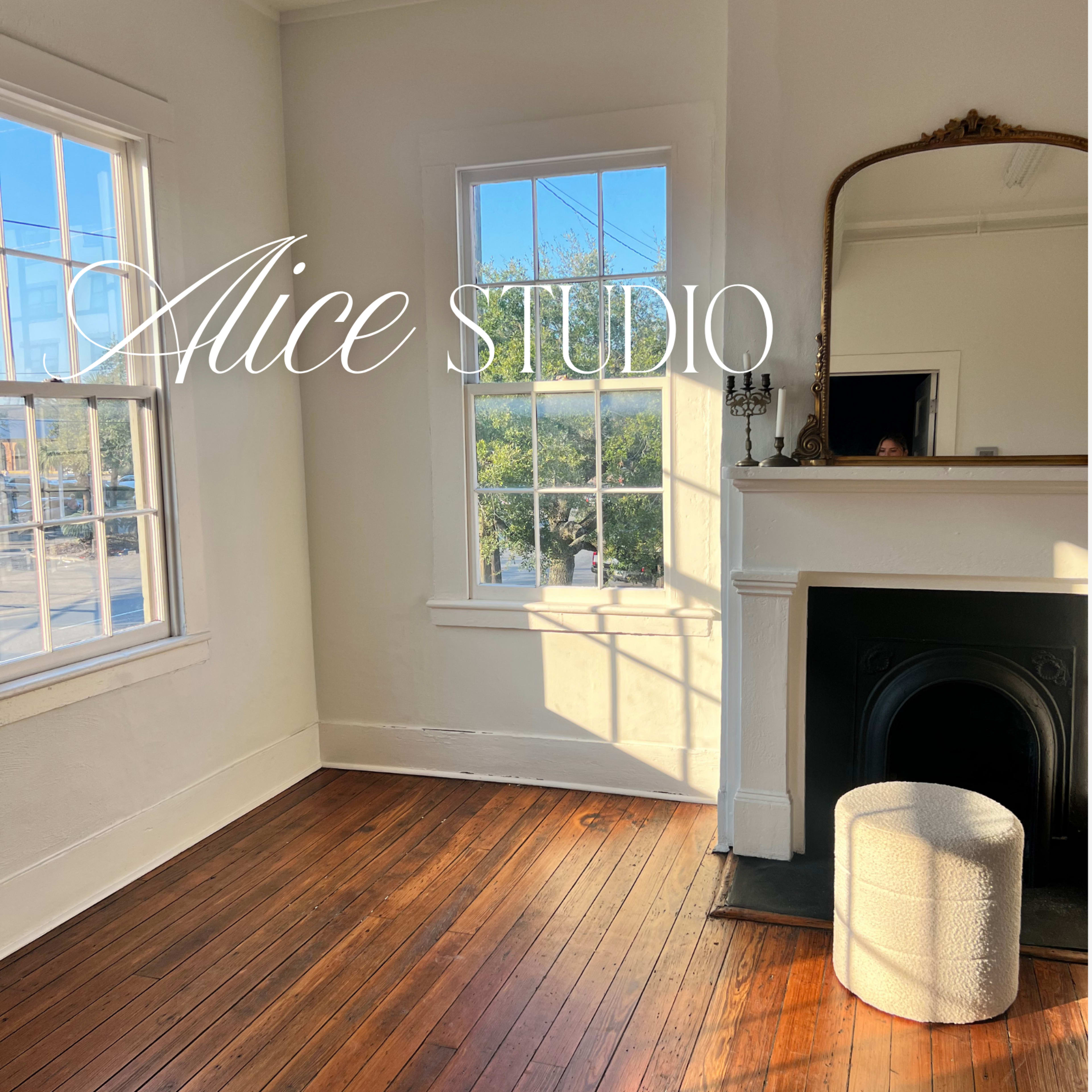 The image shows a bright, minimally furnished room with large windows, wooden flooring, and a decorative mirror above a fireplace.