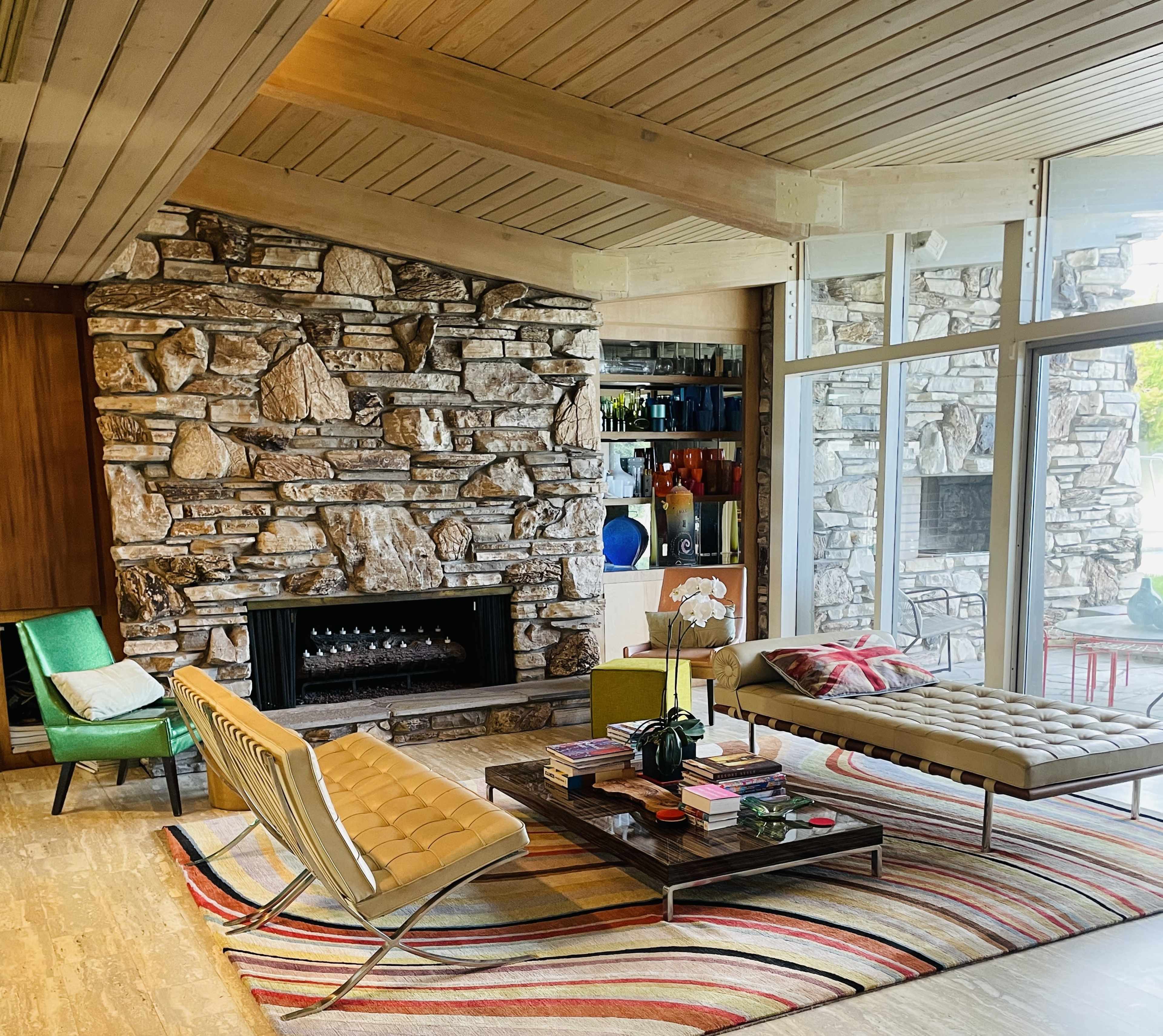 The image shows a modern living room with a stone wall, a fireplace, a coffee table, and a colorful area rug, surrounded by large windows.