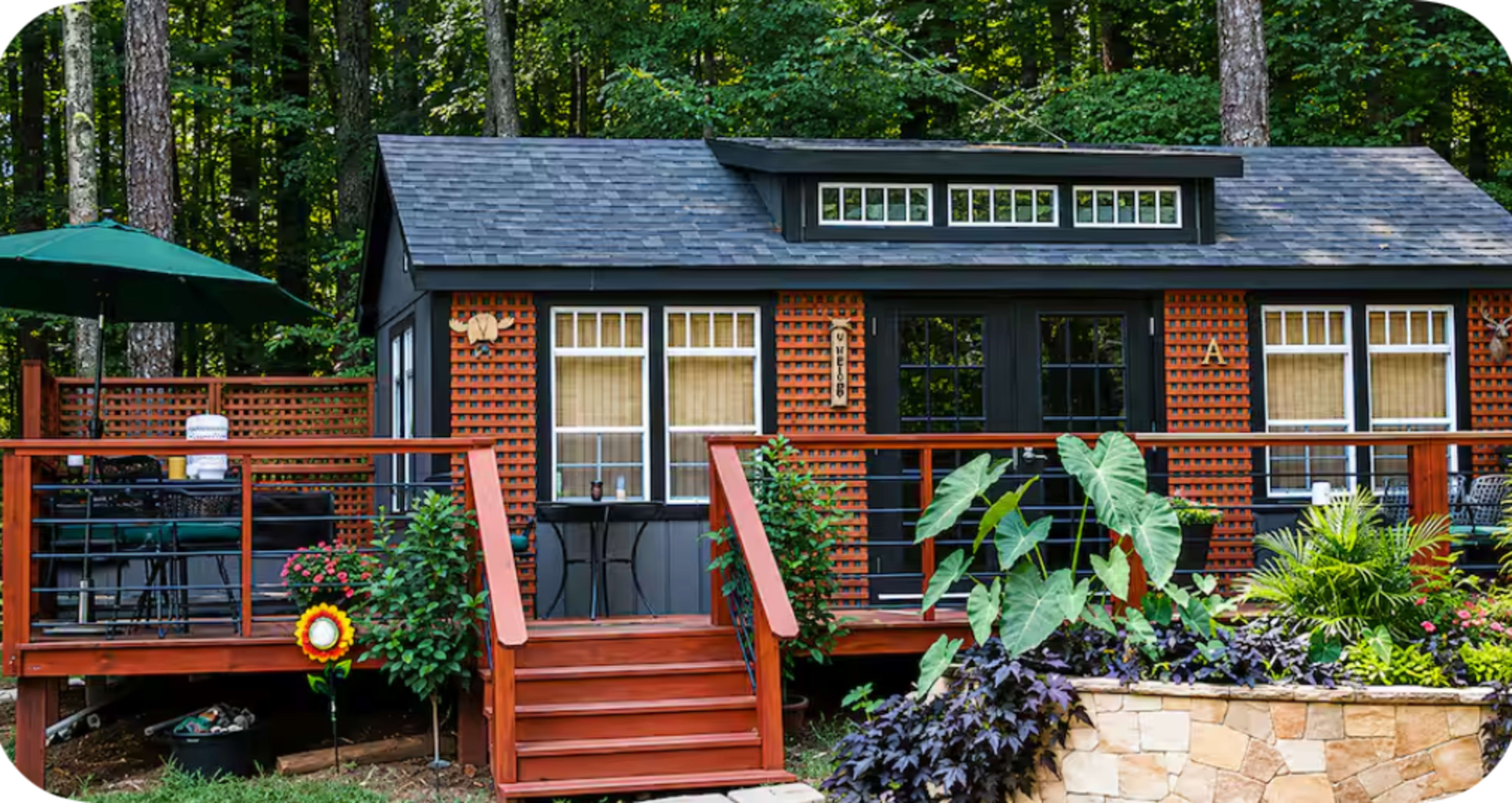A small, black cabin with wooden features and a deck is surrounded by greenery and outdoor furniture.