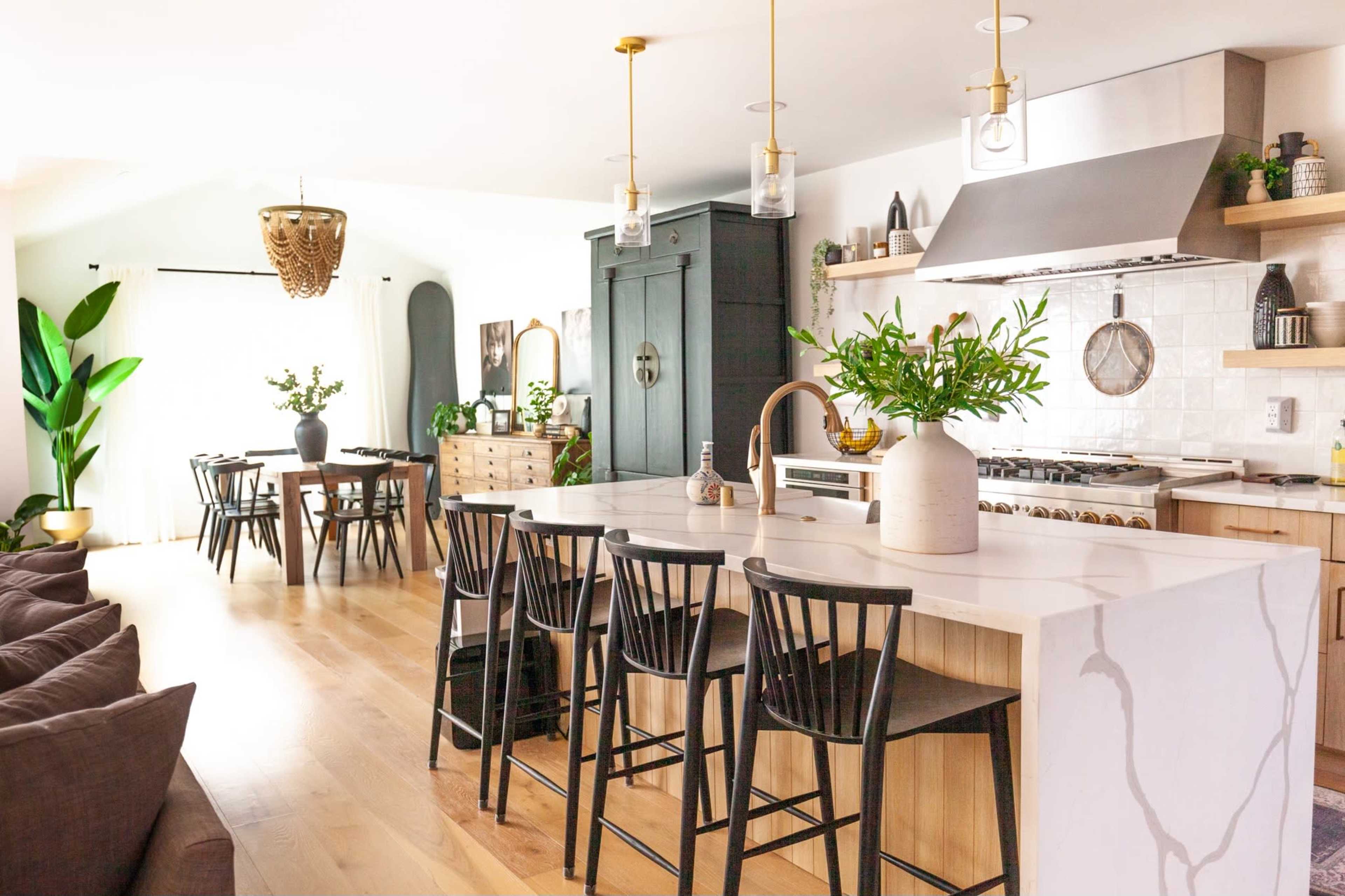 A modern kitchen with a large island featuring black stools, a dining area in the background with a wooden table, and various plants throughout the space.