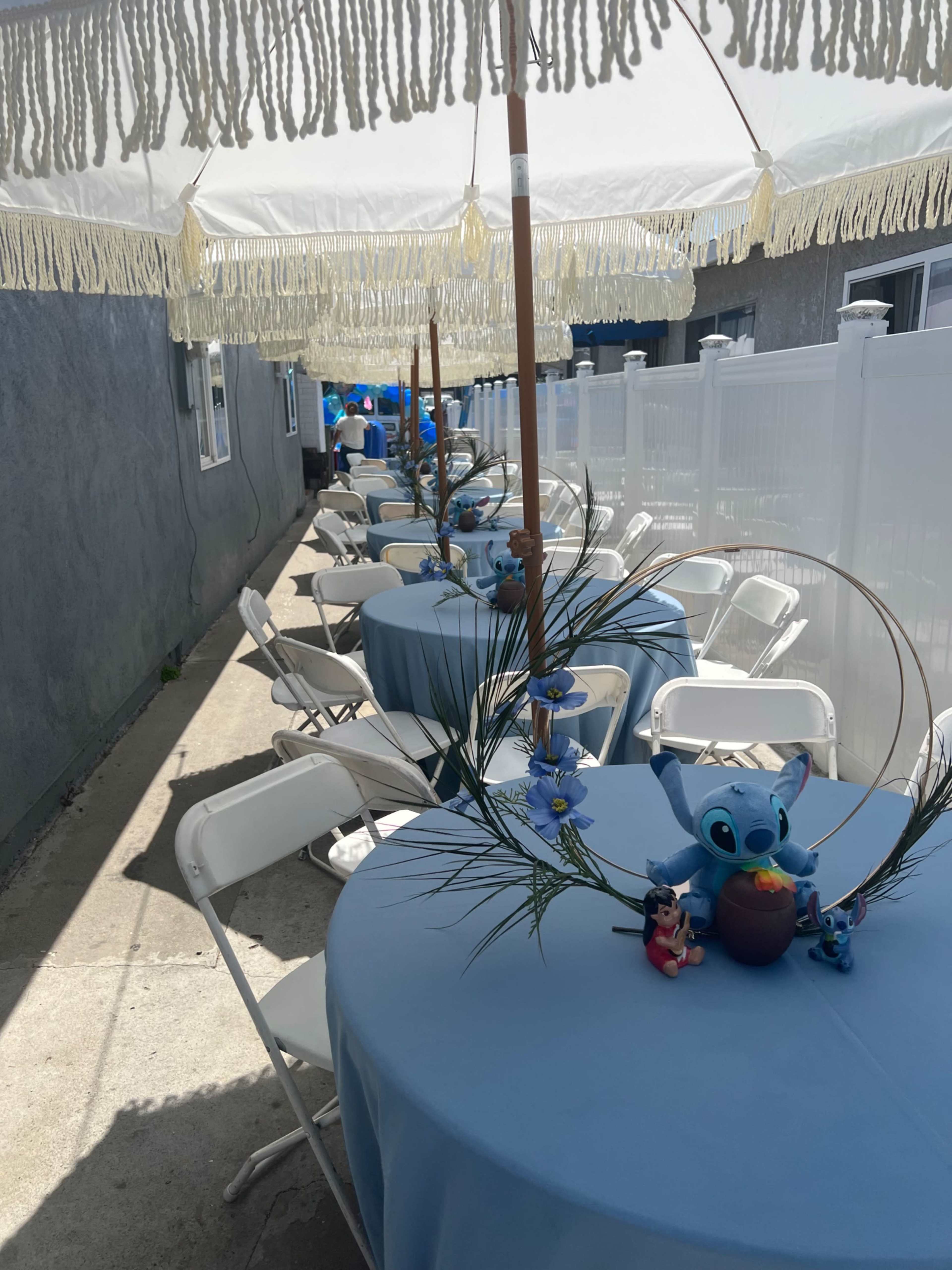 A row of tables covered in light blue tablecloths is set up outdoors under a decorative canopy, with potted plants and small figurines arranged in the center of each table.