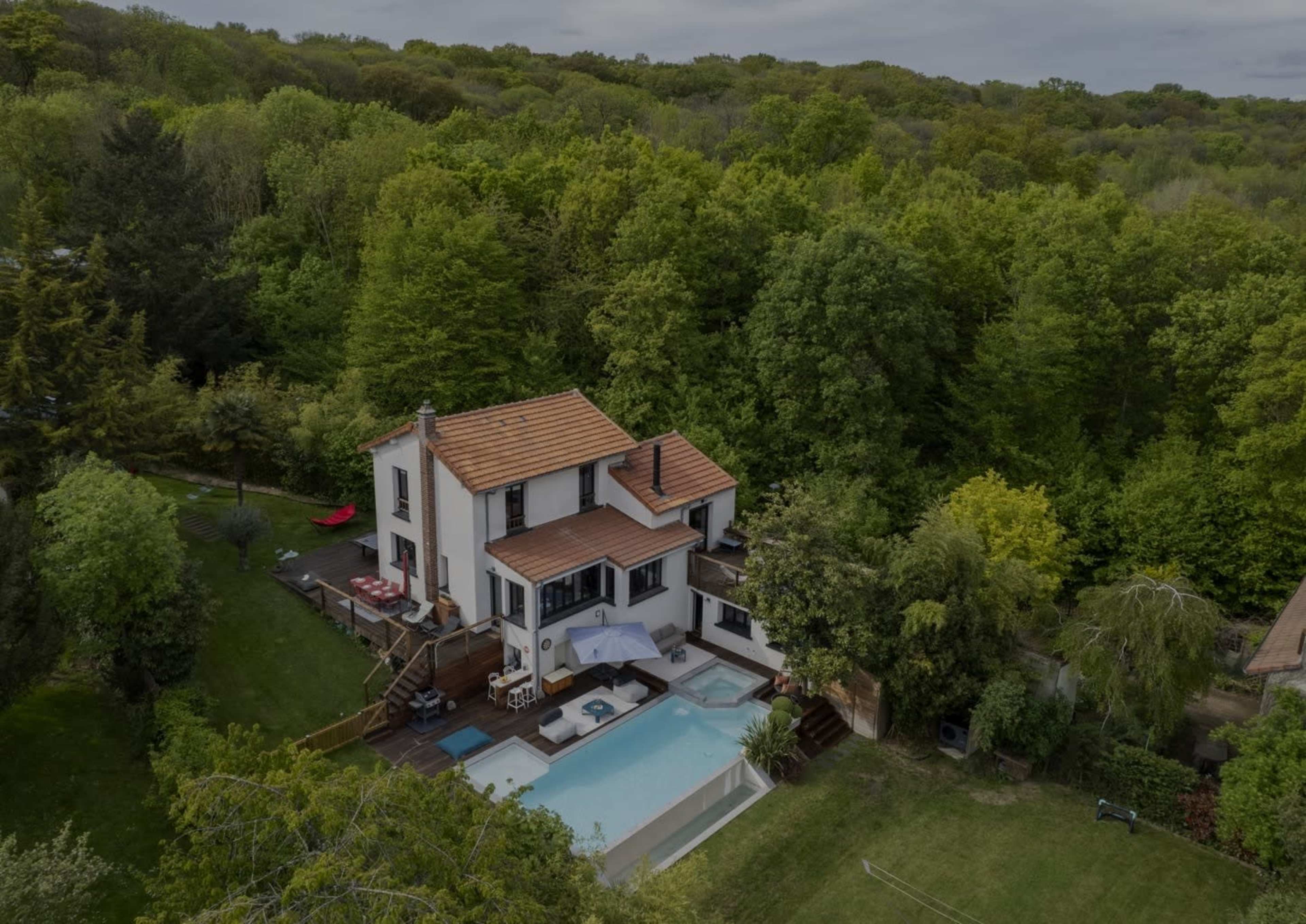 A two-story white house with a brown roof is surrounded by lush green trees and features a swimming pool in the backyard.