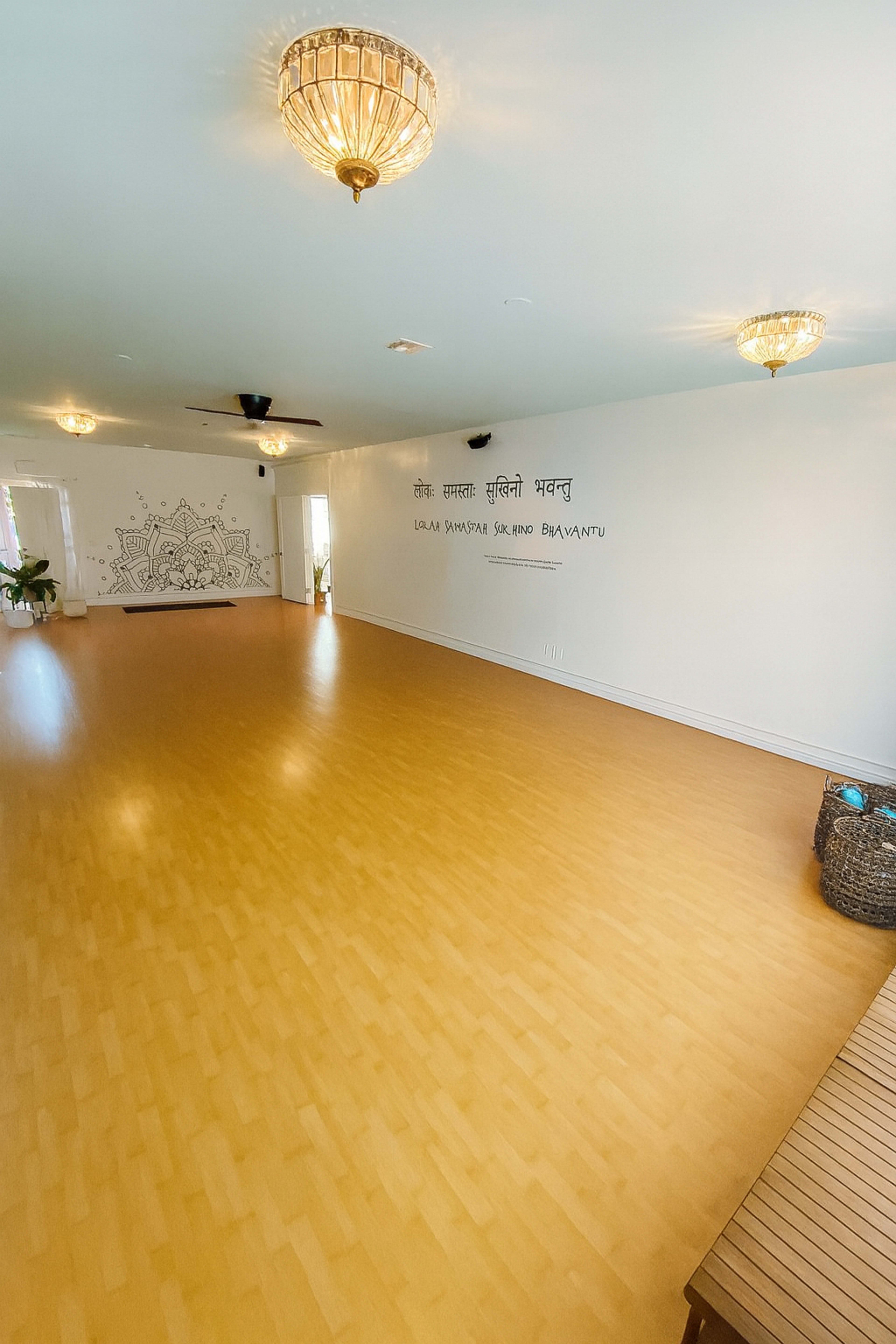 The image shows a spacious and empty yoga studio with wooden flooring and decorative lighting fixtures.