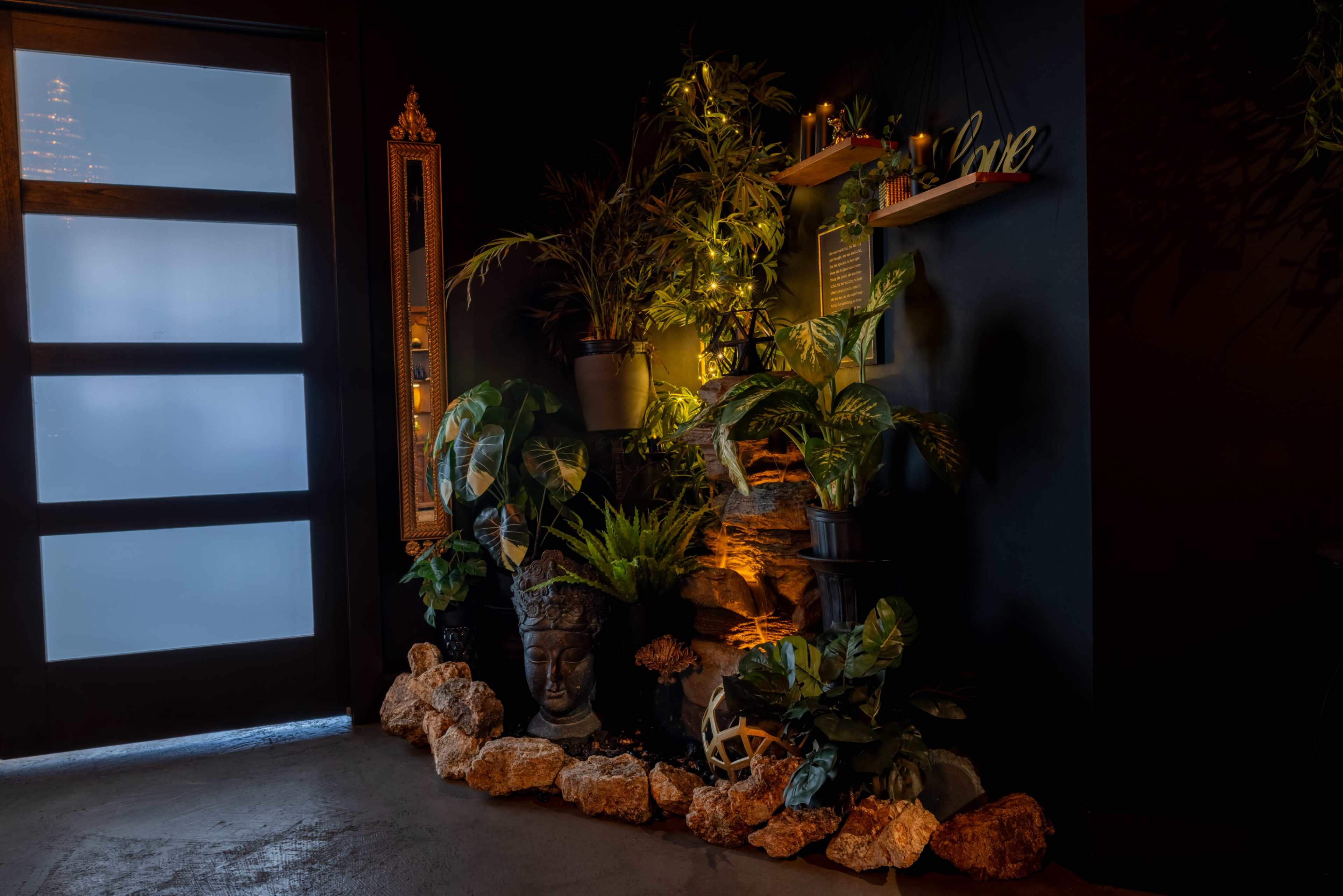 A corner featuring a collection of indoor plants, a decorative stone arrangement, and a large Buddha statue, illuminated by soft light against a dark wall.