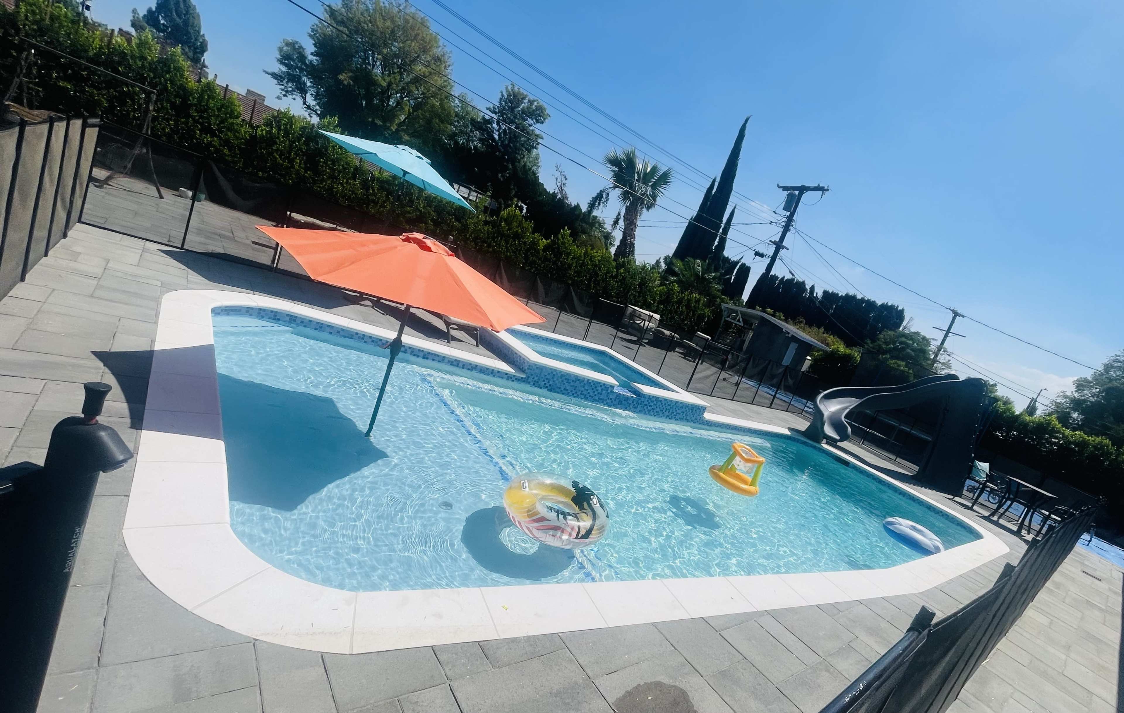 The image shows a swimming pool with a water slide, an orange umbrella, and a floating lounge in a fenced yard.