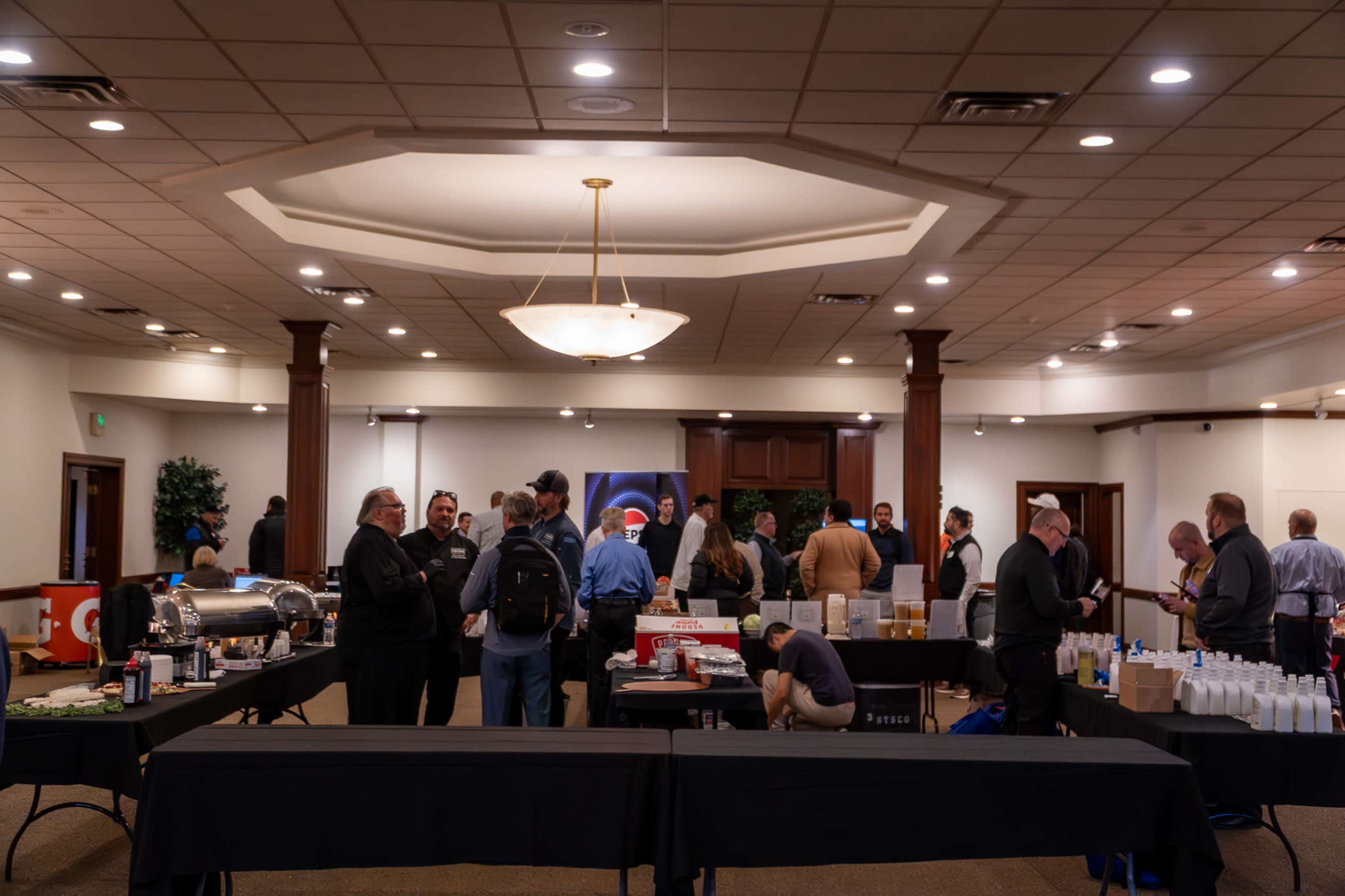 A group of people gathers in a spacious indoor venue, engaging in conversation while tables are set up with various food and drink items.