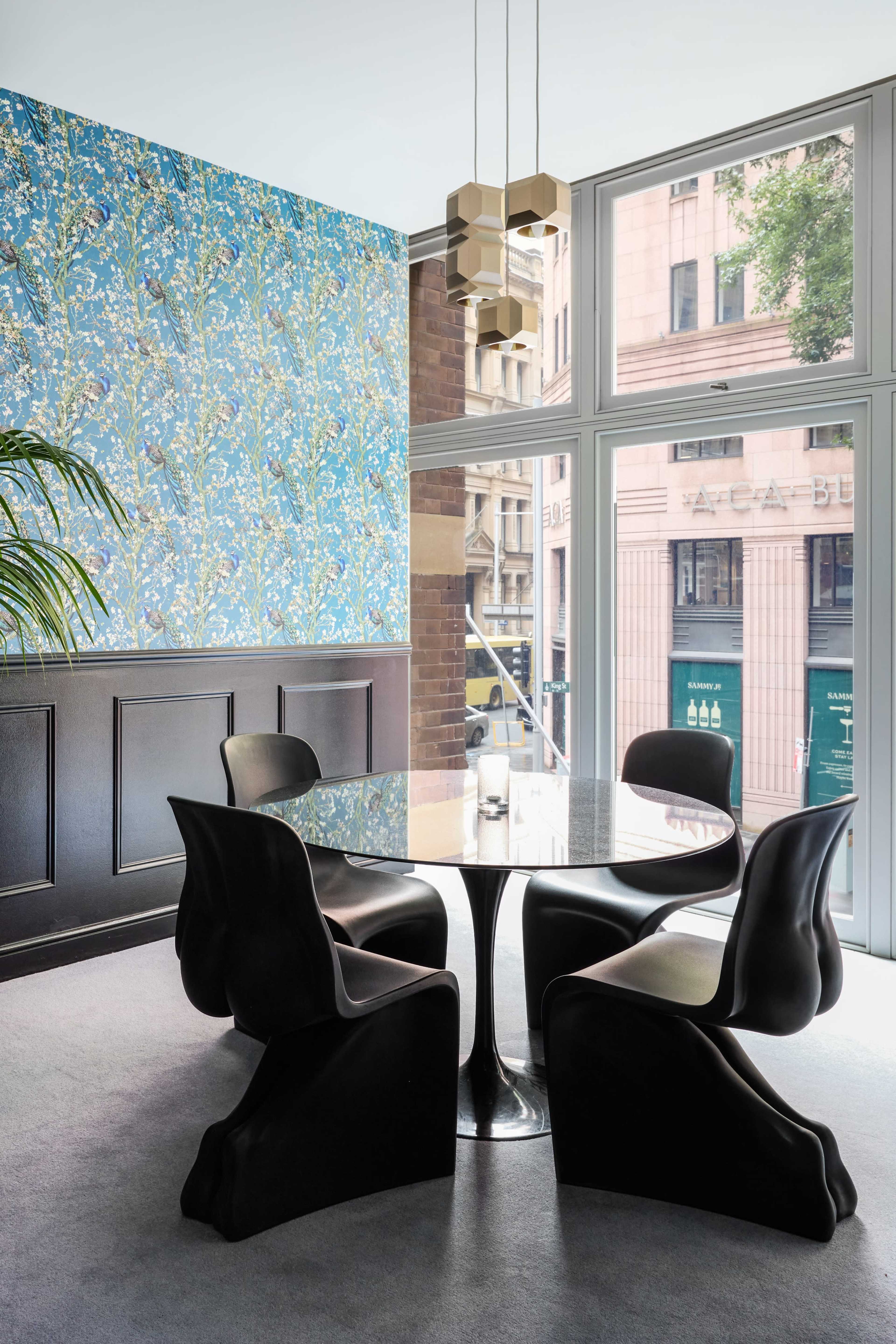 A modern dining area features four black chairs around a round glass table, set against a blue floral wallpaper and large windows with a city view.