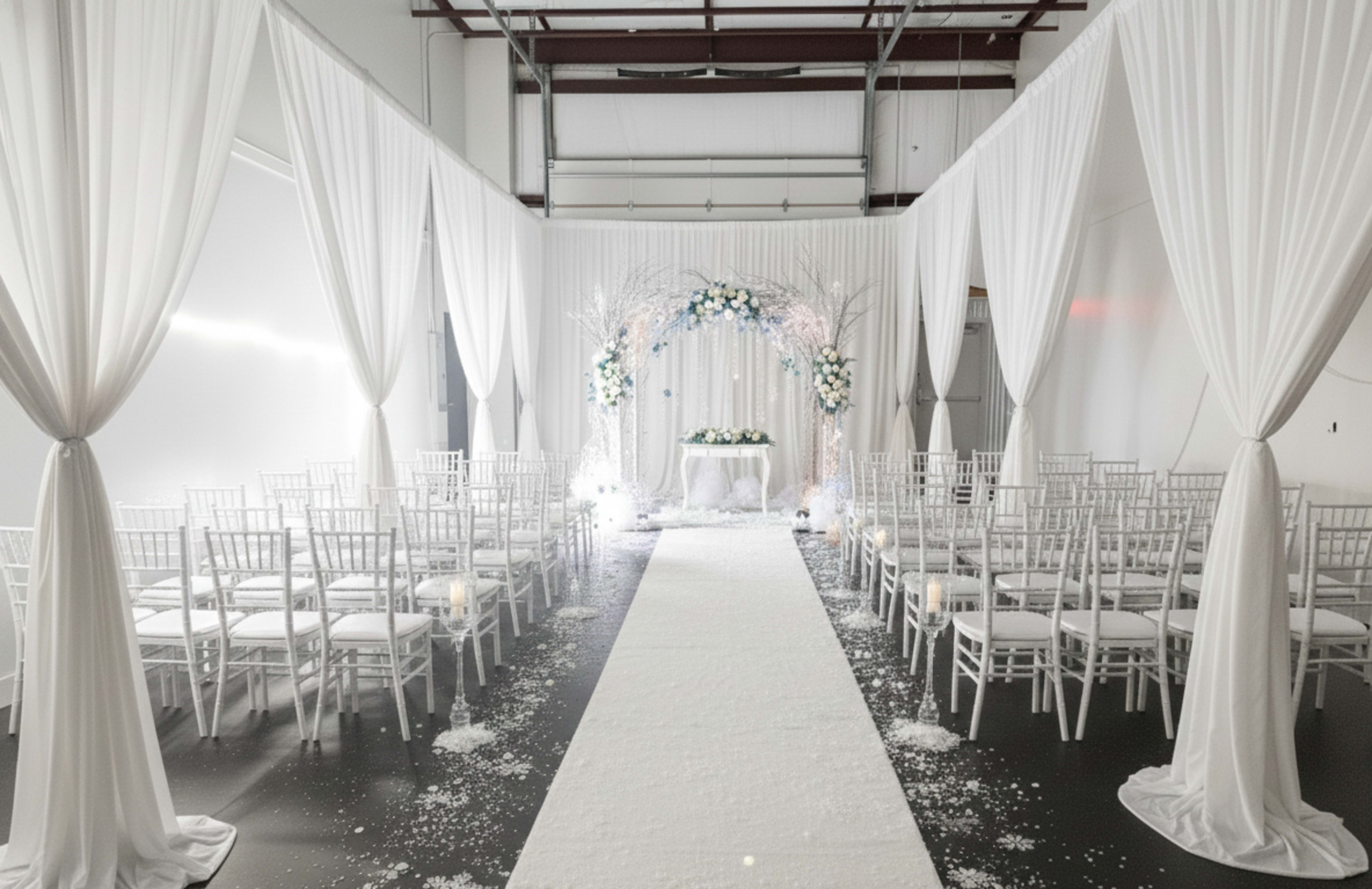 The image shows a wedding ceremony setup featuring white draped fabric, rows of white chairs, and a floral arch at the front on a carpeted aisle.