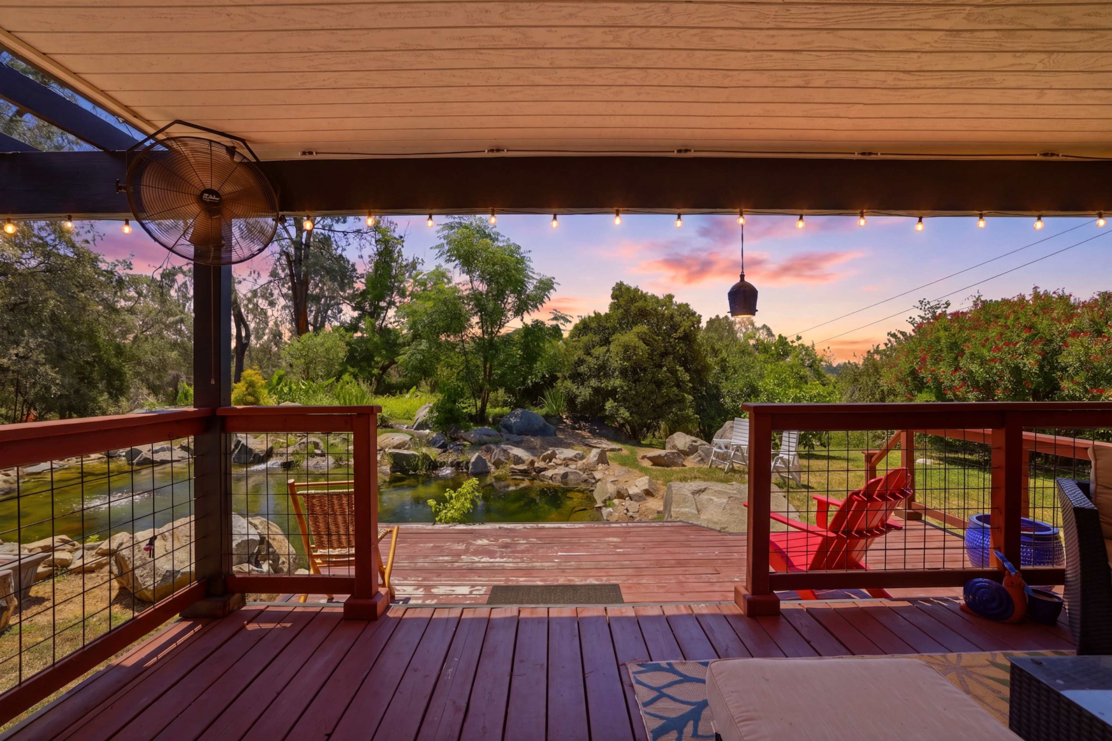 The image shows a wooden deck overlooking a tranquil pond with rocks and greenery, framed by a colorful sunset sky.