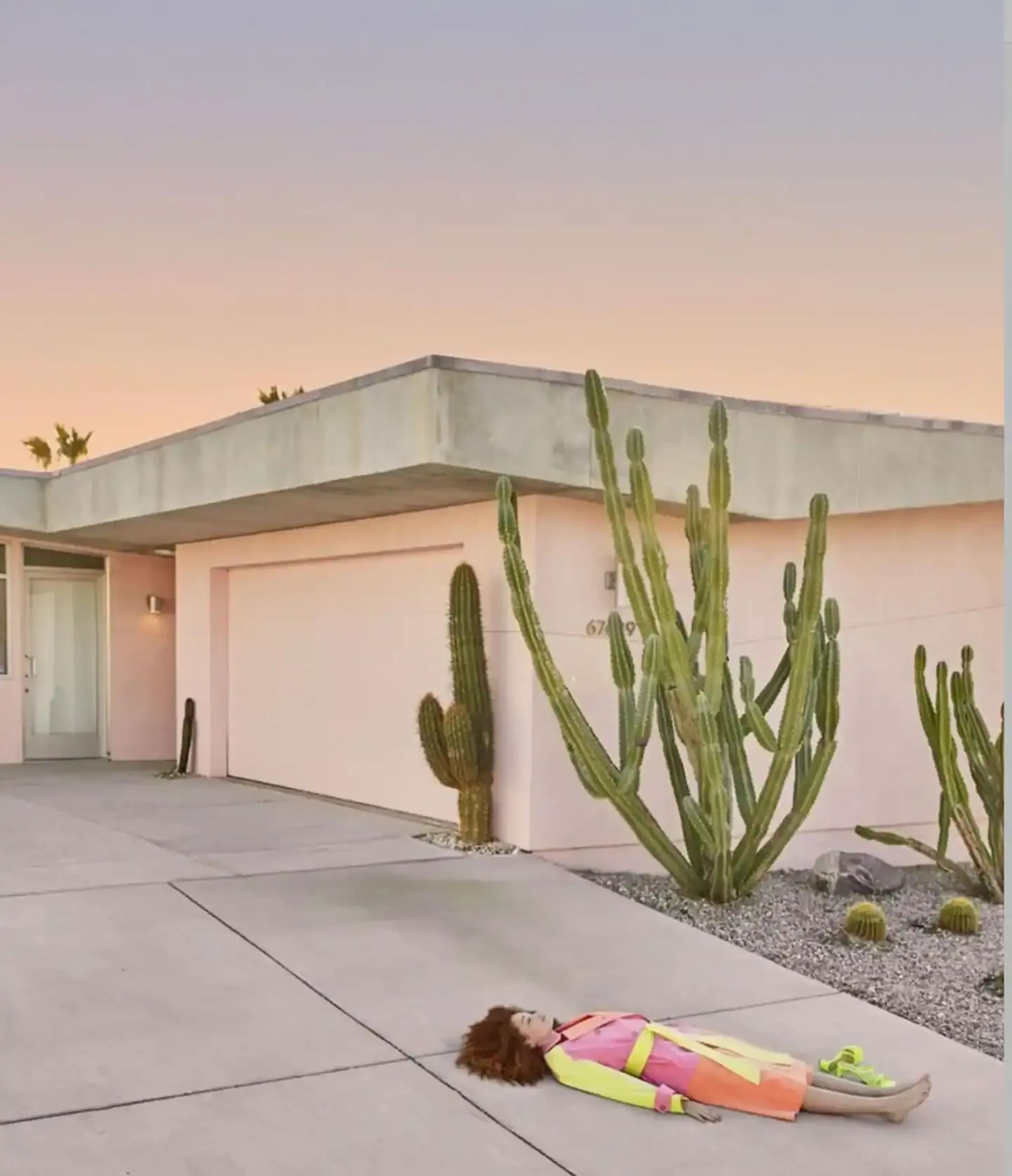 A woman lies on the concrete driveway of a pink house surrounded by tall cacti under a gradient sky.