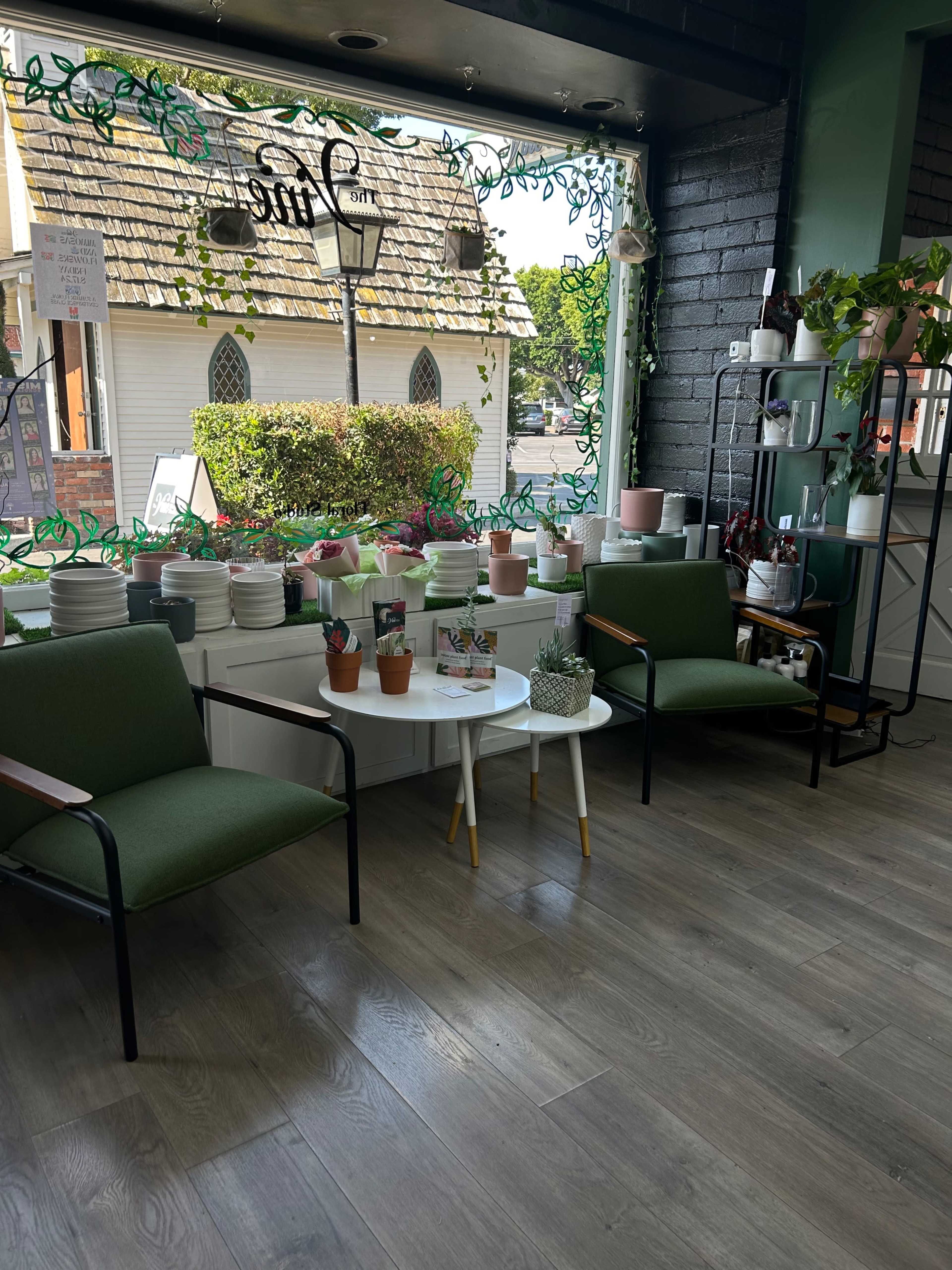The image shows a cozy café interior with two green chairs, a white table, and a display of potted plants and dishware near a window.