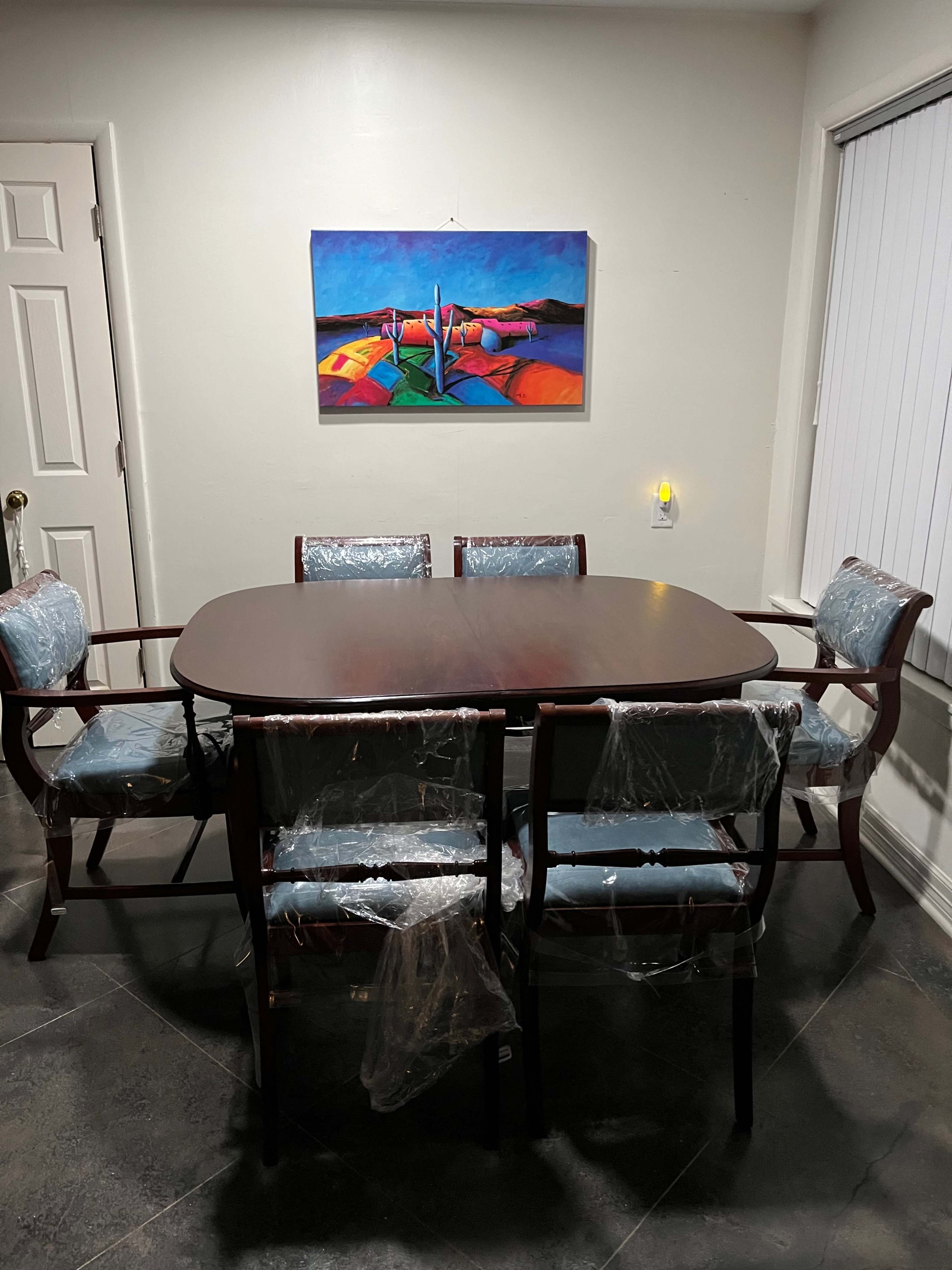 A wooden dining table is surrounded by six chairs covered in plastic, under a colorful painting on the wall.