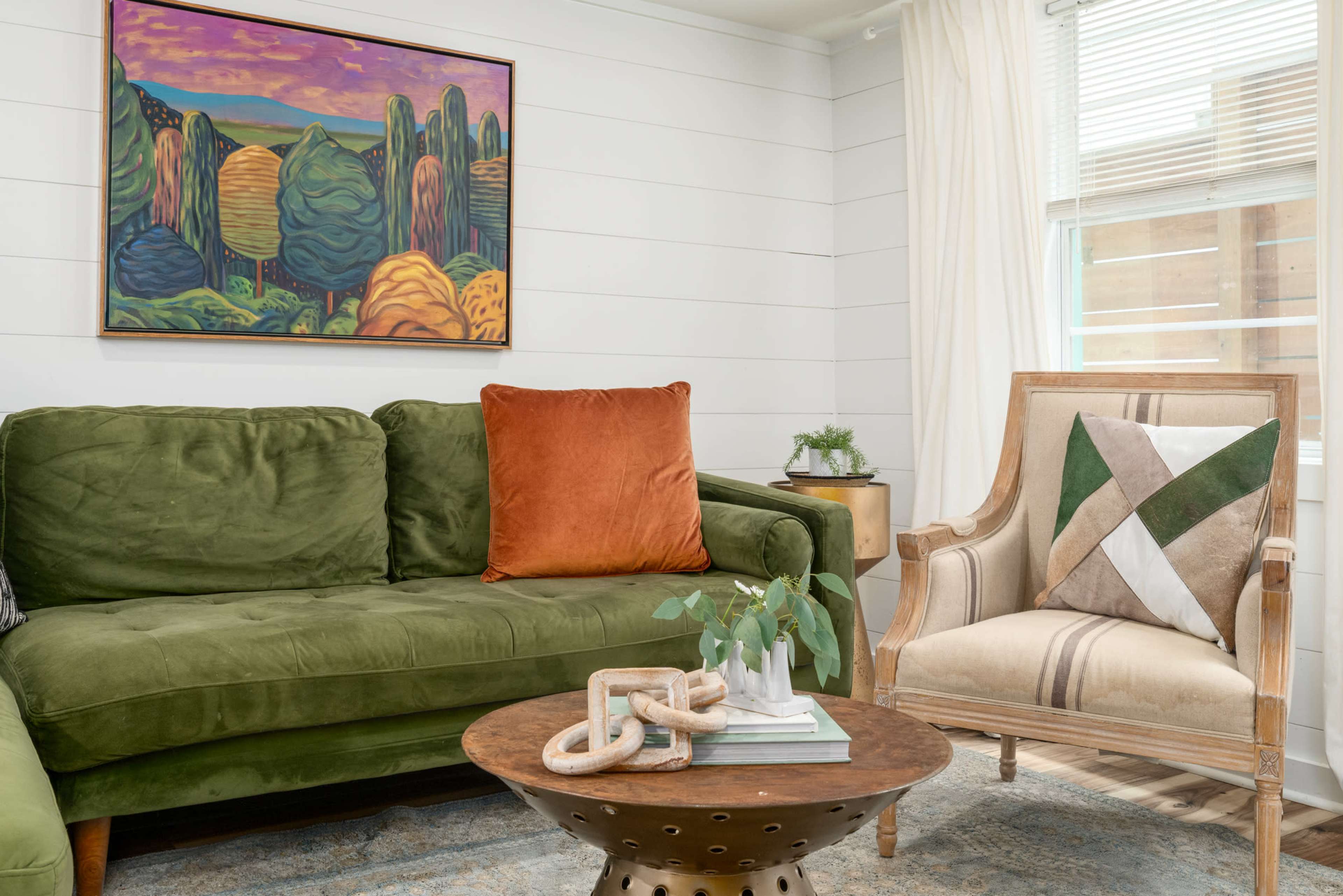 A green sofa with an orange pillow and a patterned chair are positioned around a wooden coffee table, with a colorful landscape painting hanging on the wall.