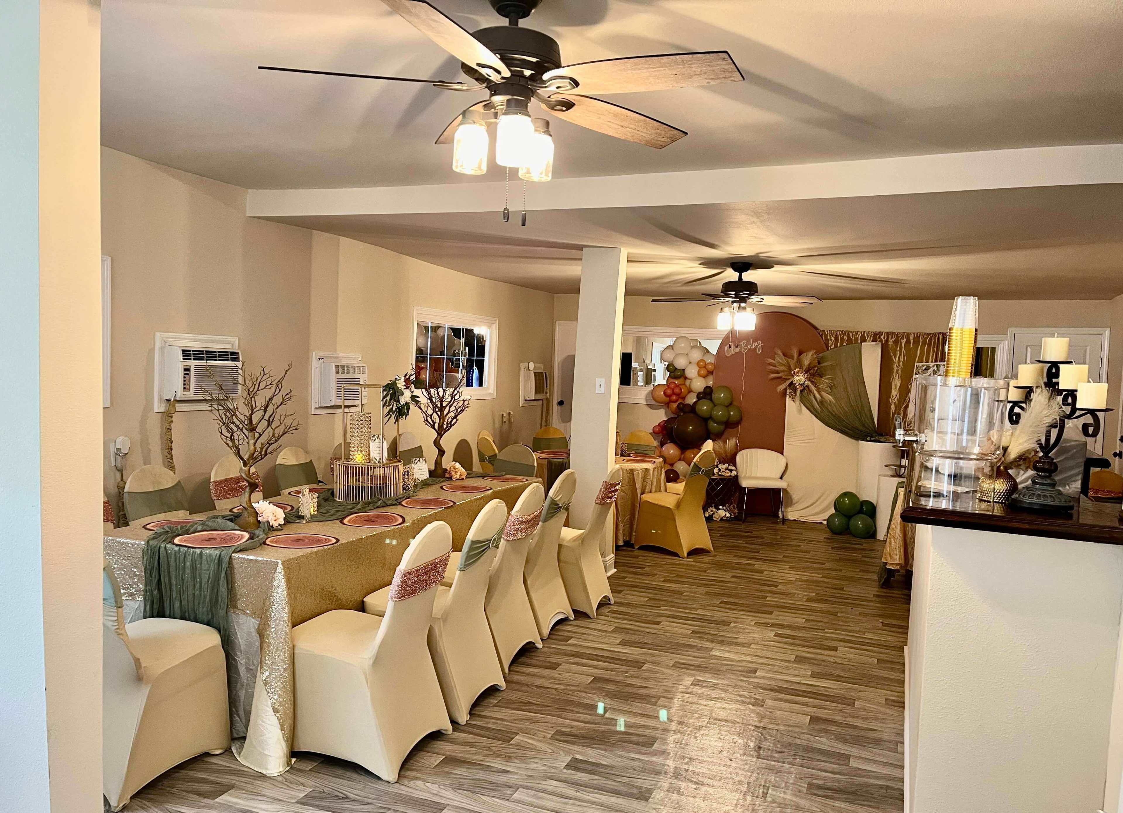The image depicts a decorated dining area with a long table set for a gathering, featuring elegant tableware and a backdrop of balloons and greenery.