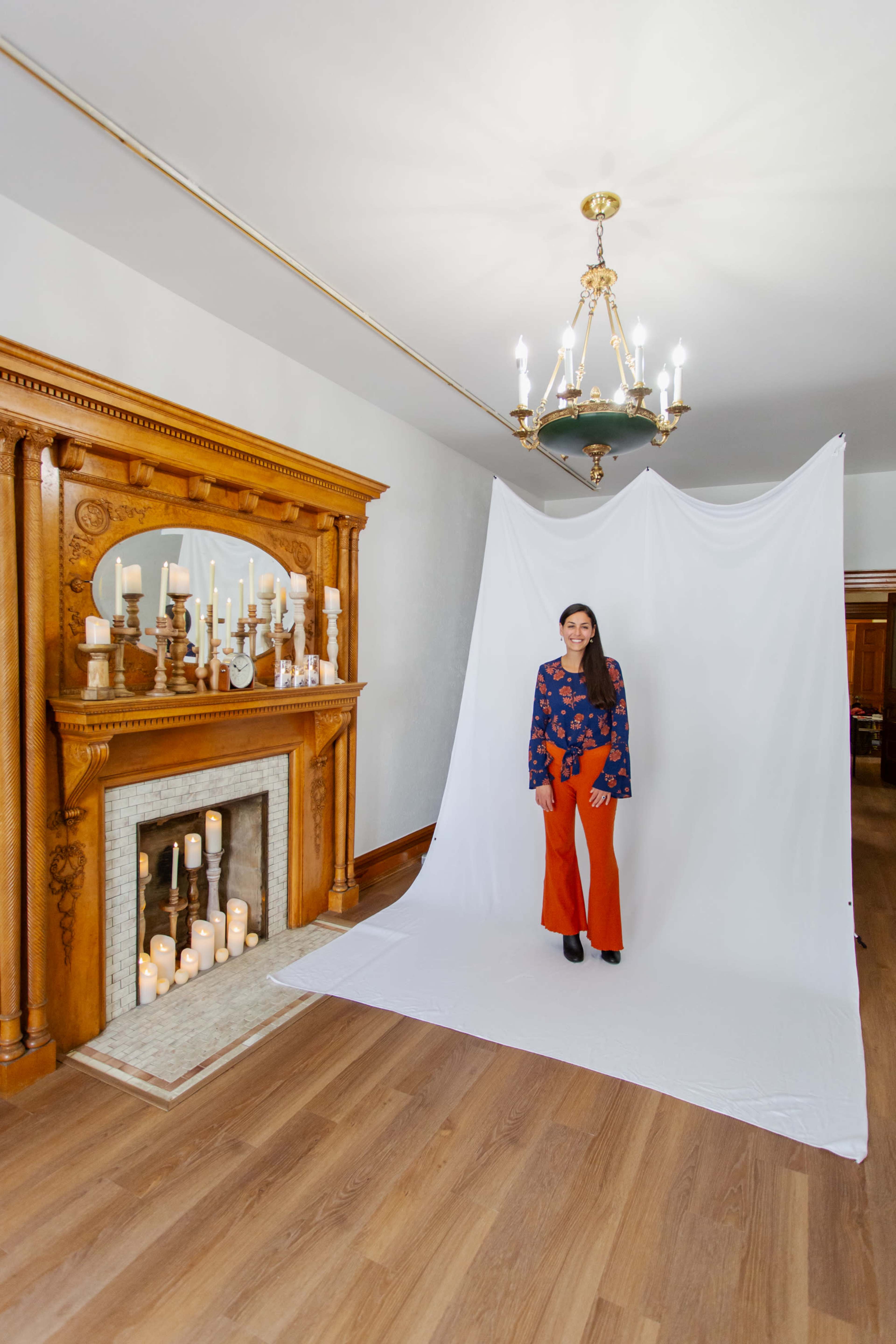 A woman stands on a white backdrop in a room featuring a decorative fireplace with candles and a chandelier.