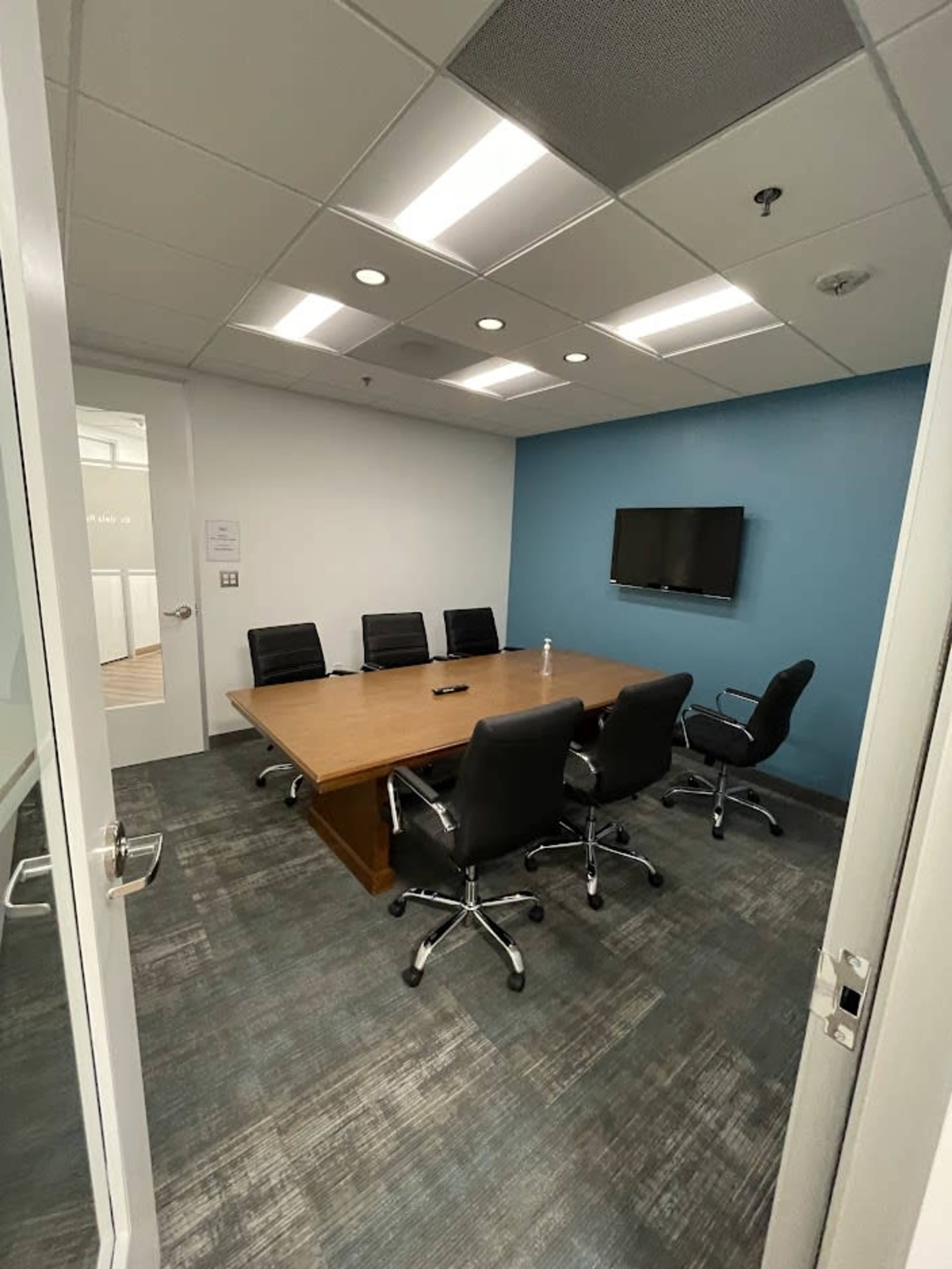 A meeting room features a large wooden table surrounded by black chairs, with a wall-mounted TV and blue accent wall.