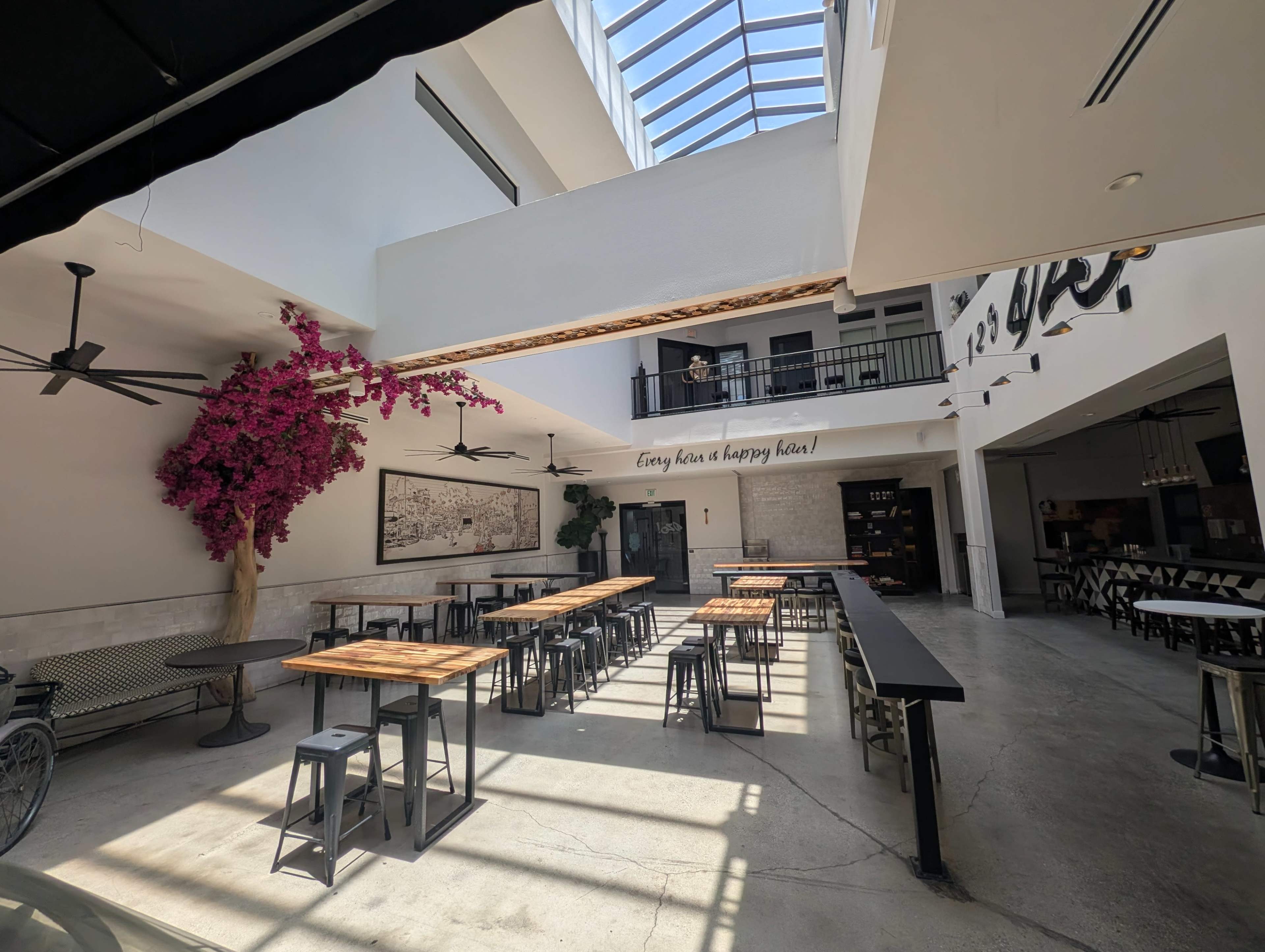 The image shows a spacious, bright café with wooden tables, metal stools, and a large pink tree near the entrance, all under a skylight.