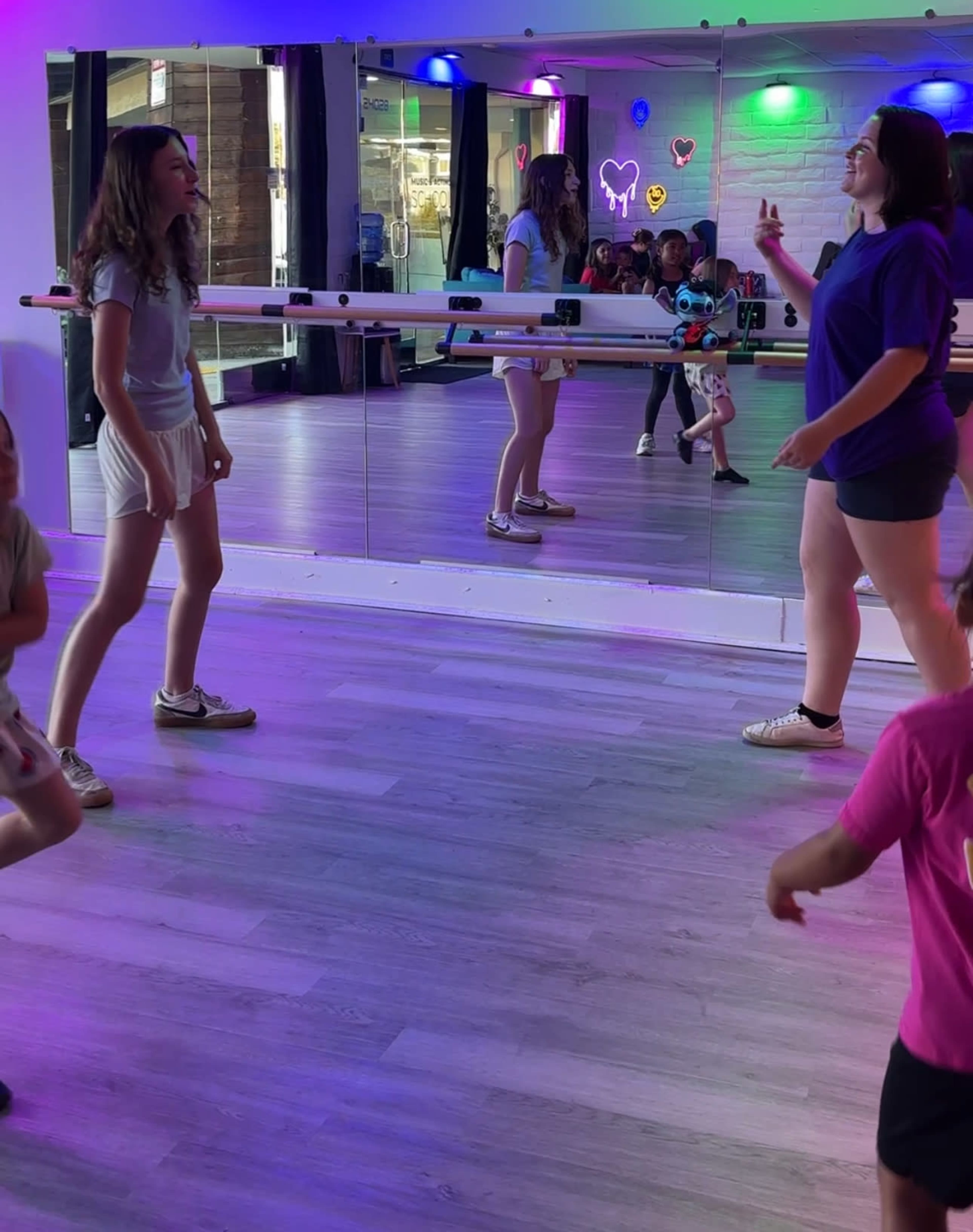A group of children and adults are engaged in a dance activity inside a brightly lit studio with mirrors and colorful lighting.