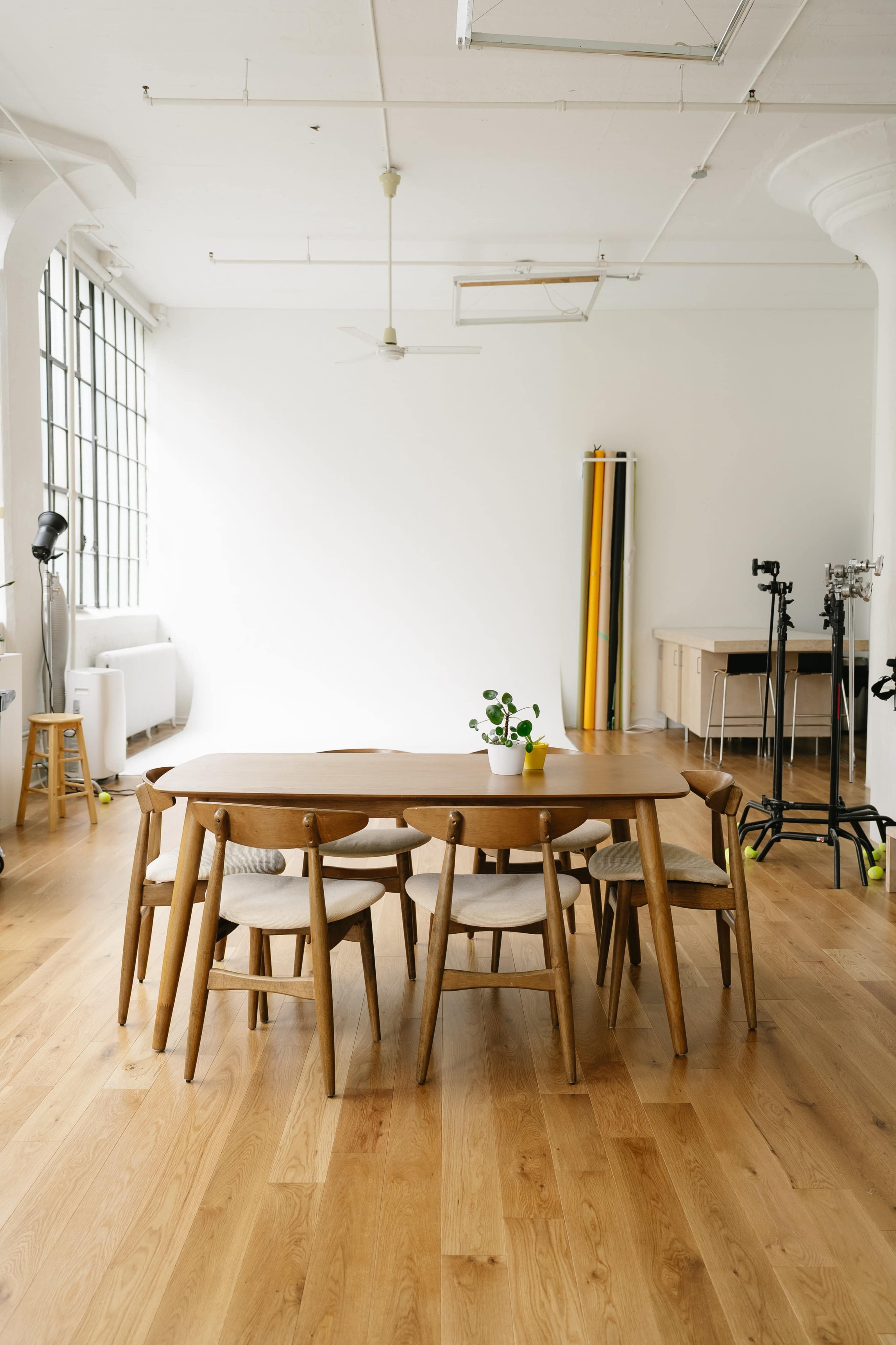 A large wooden table surrounded by several chairs is positioned in a spacious, well-lit room with minimal decor and a simple color palette.