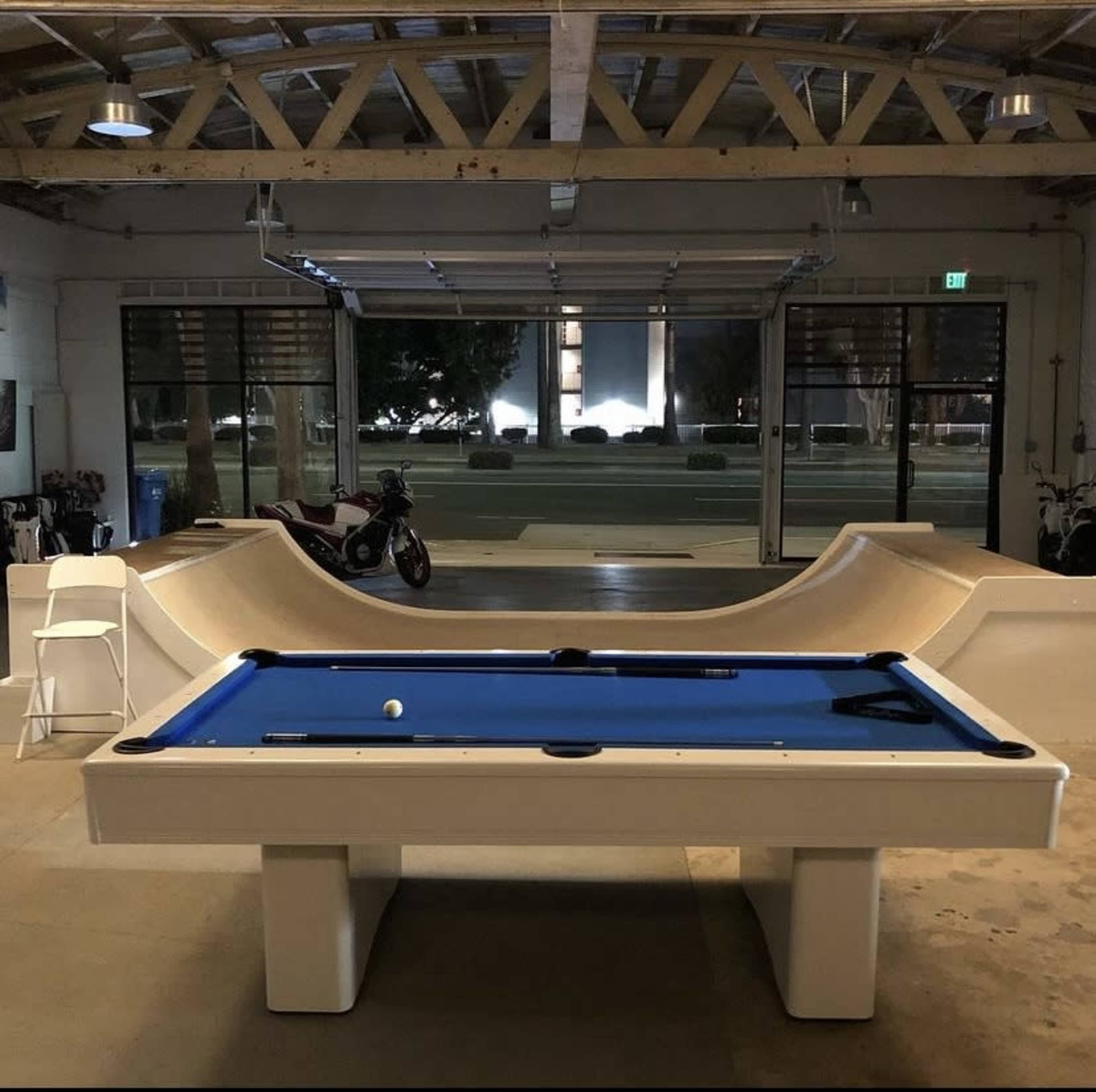 A blue pool table is positioned in an industrial-style space with large windows and a skate ramp nearby.