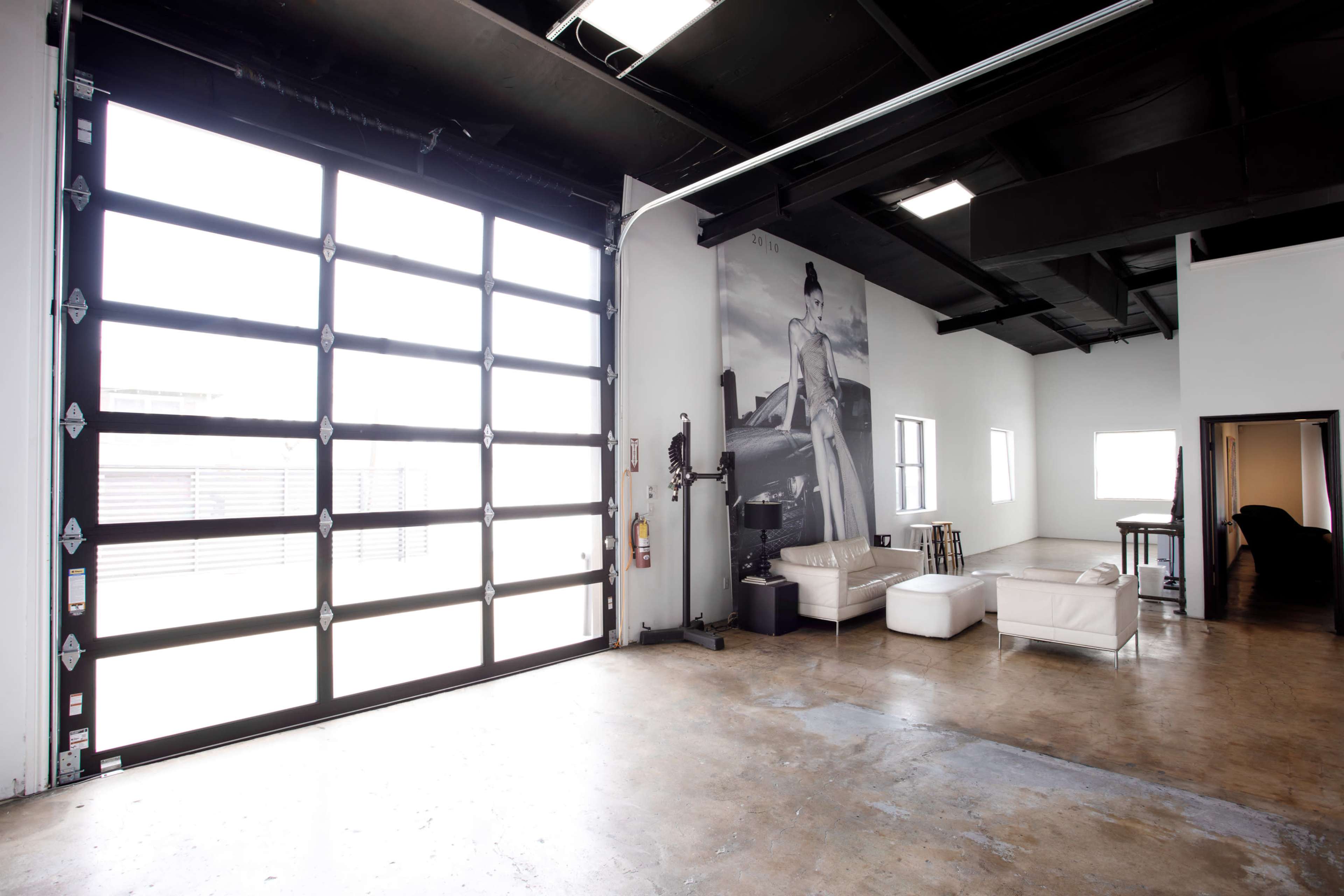 A spacious, open room with large windows, a glass garage door, and minimal furniture, including white sofas and a table.