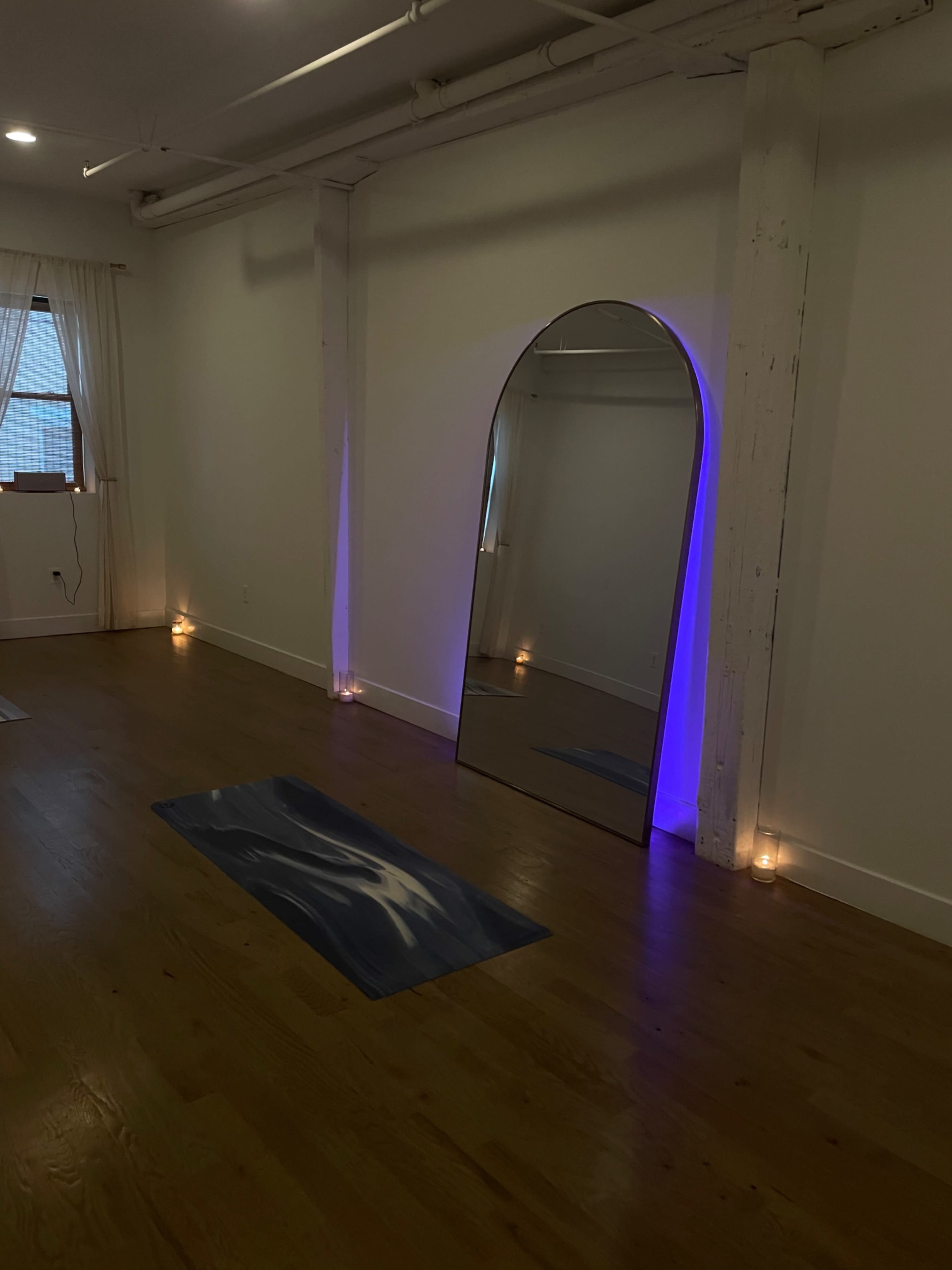 The image shows a yoga studio with a large mirror framed by purple lighting and yoga mats placed on a wooden floor.