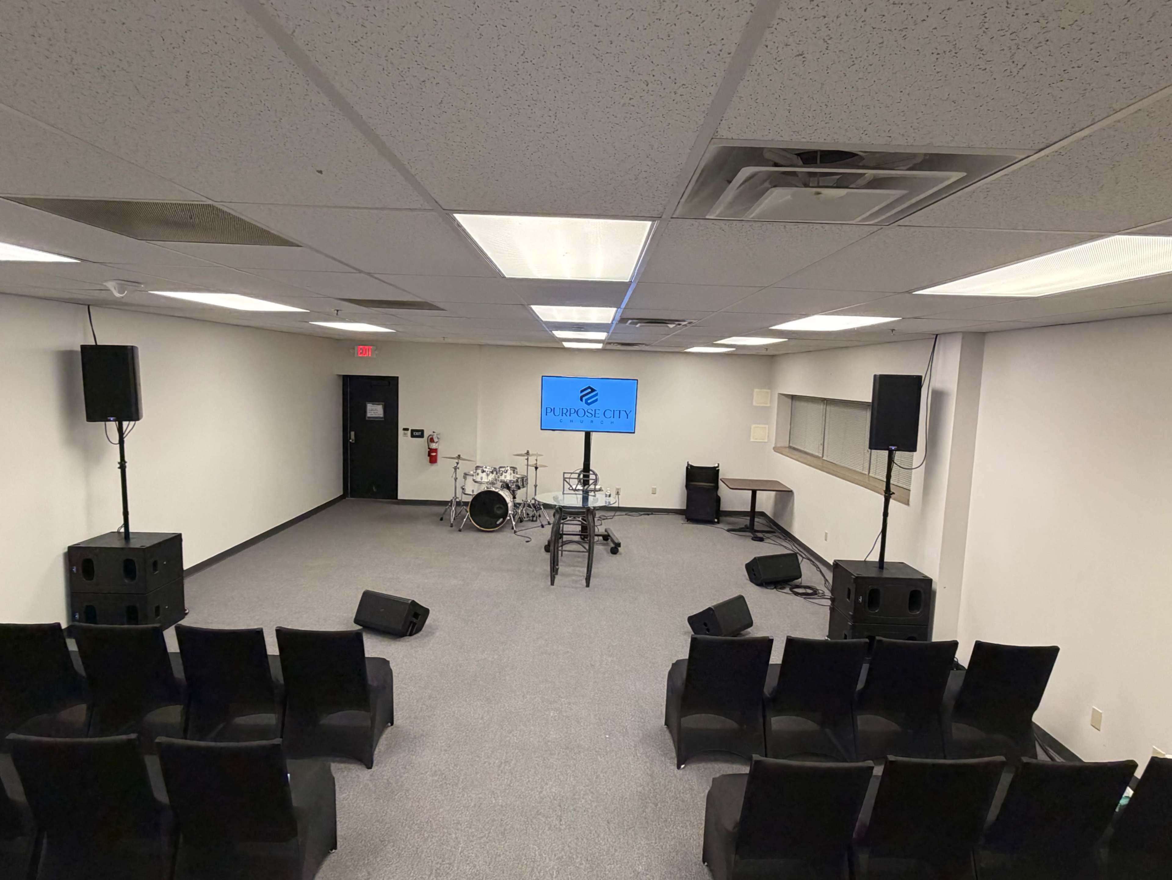 The image shows a small stage setup with musical instruments and audio equipment in a sparsely furnished room with chairs arranged for an audience.