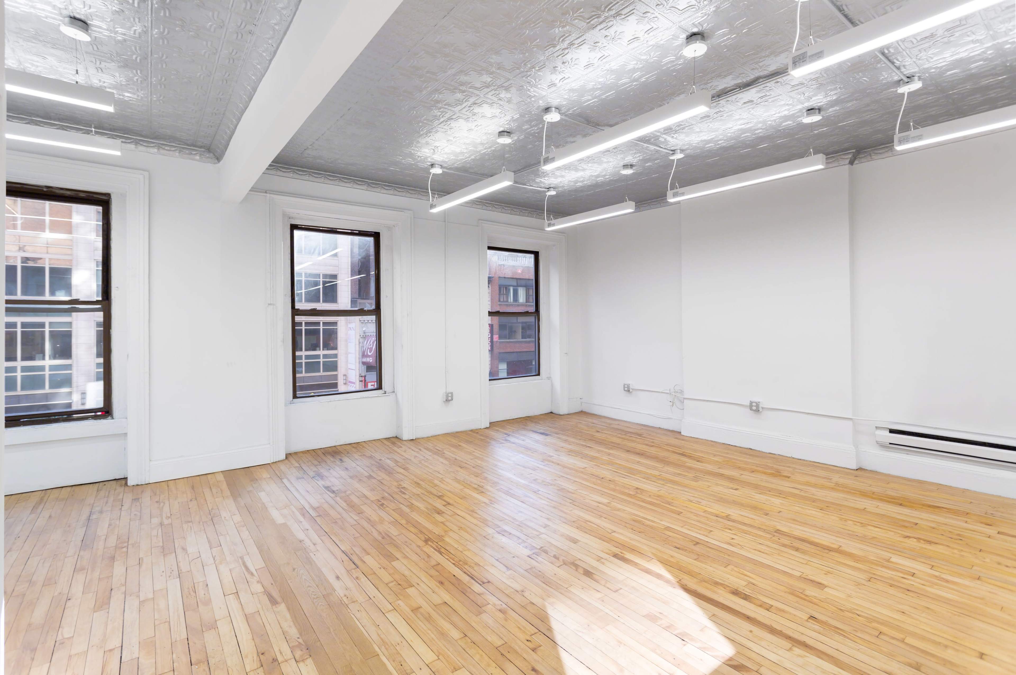 The image shows an empty room with wooden floors, large windows, and a silver tin ceiling illuminated by linear light fixtures.