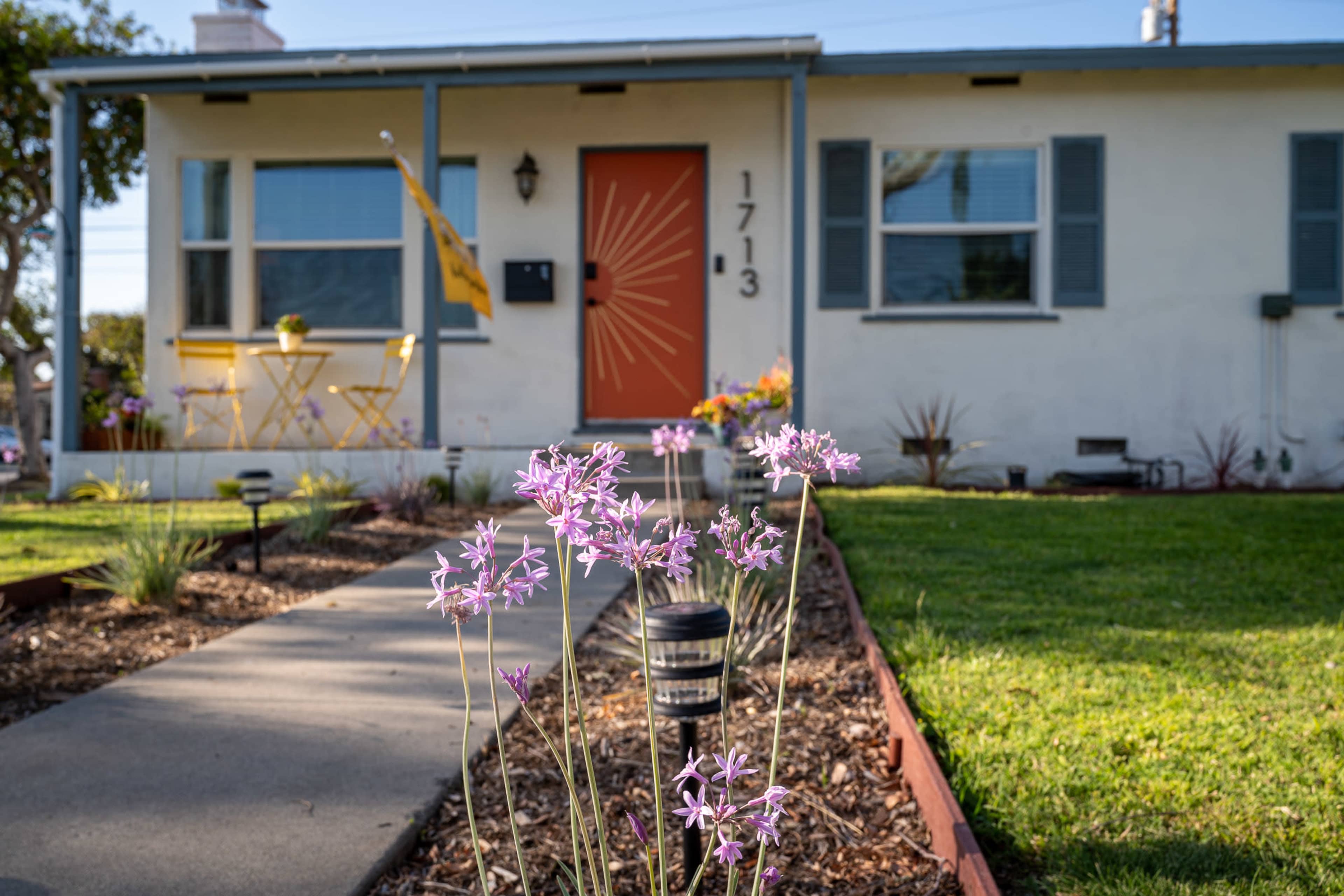A path lined with purple flowers leads to a house with a bright orange door and a small yellow bistro set on the porch.