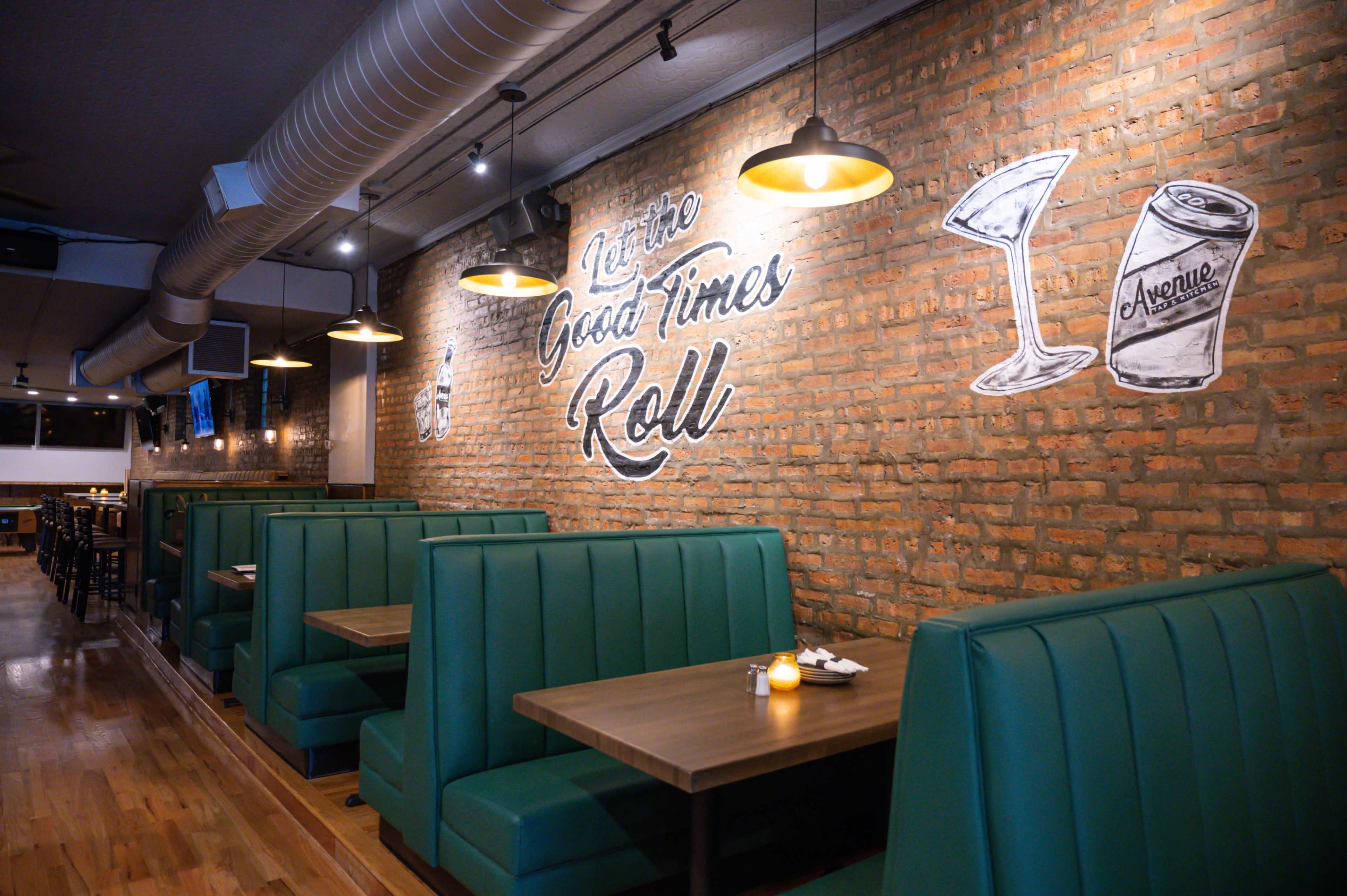 The image shows a well-lit restaurant interior with green booths and a brick wall featuring the phrase "Good Times Roll" along with illustrations of a martini glass and a can.