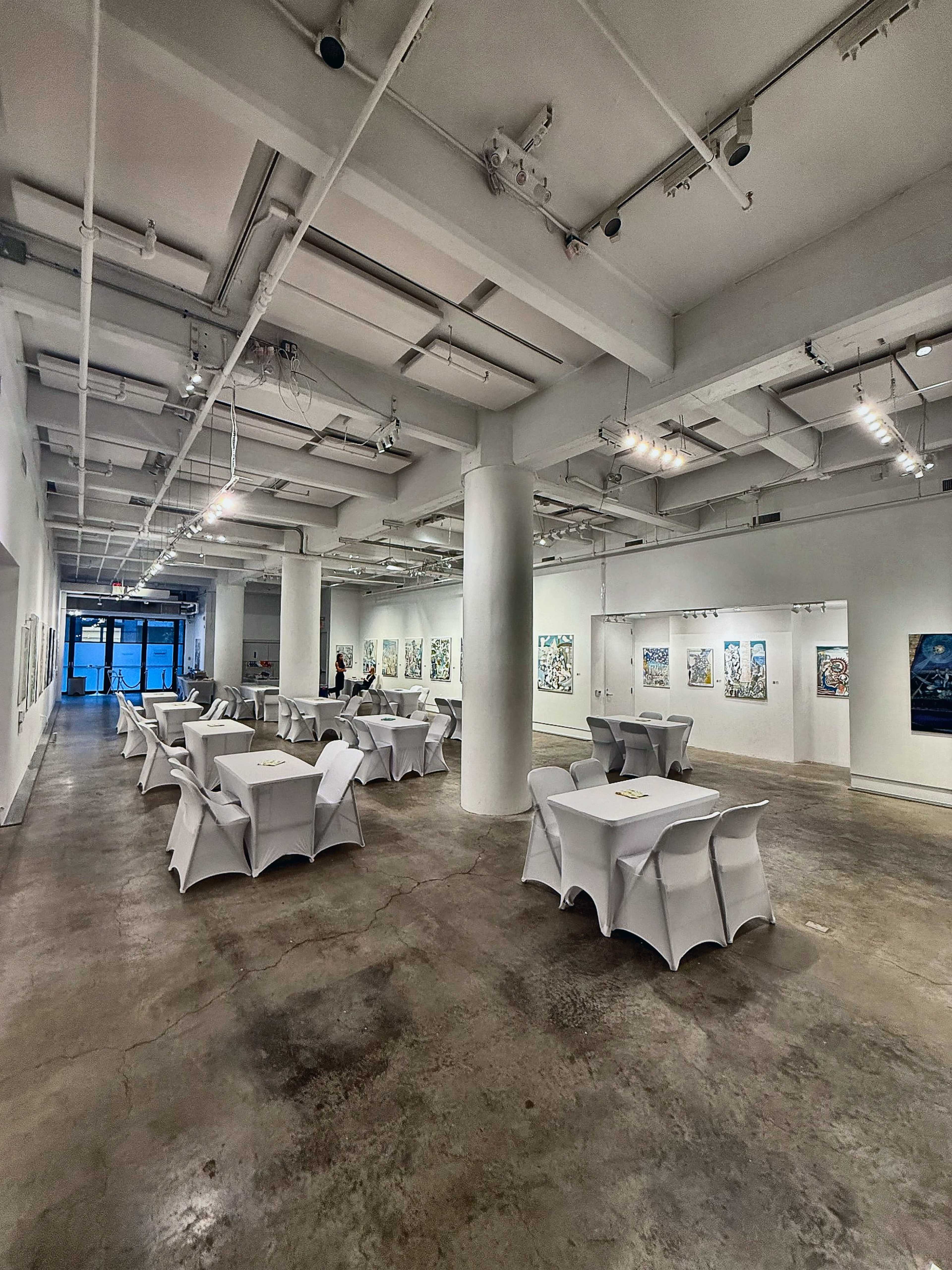 The image shows a contemporary art gallery with white walls, polished concrete floors, and tables covered in white cloth arranged for an event.