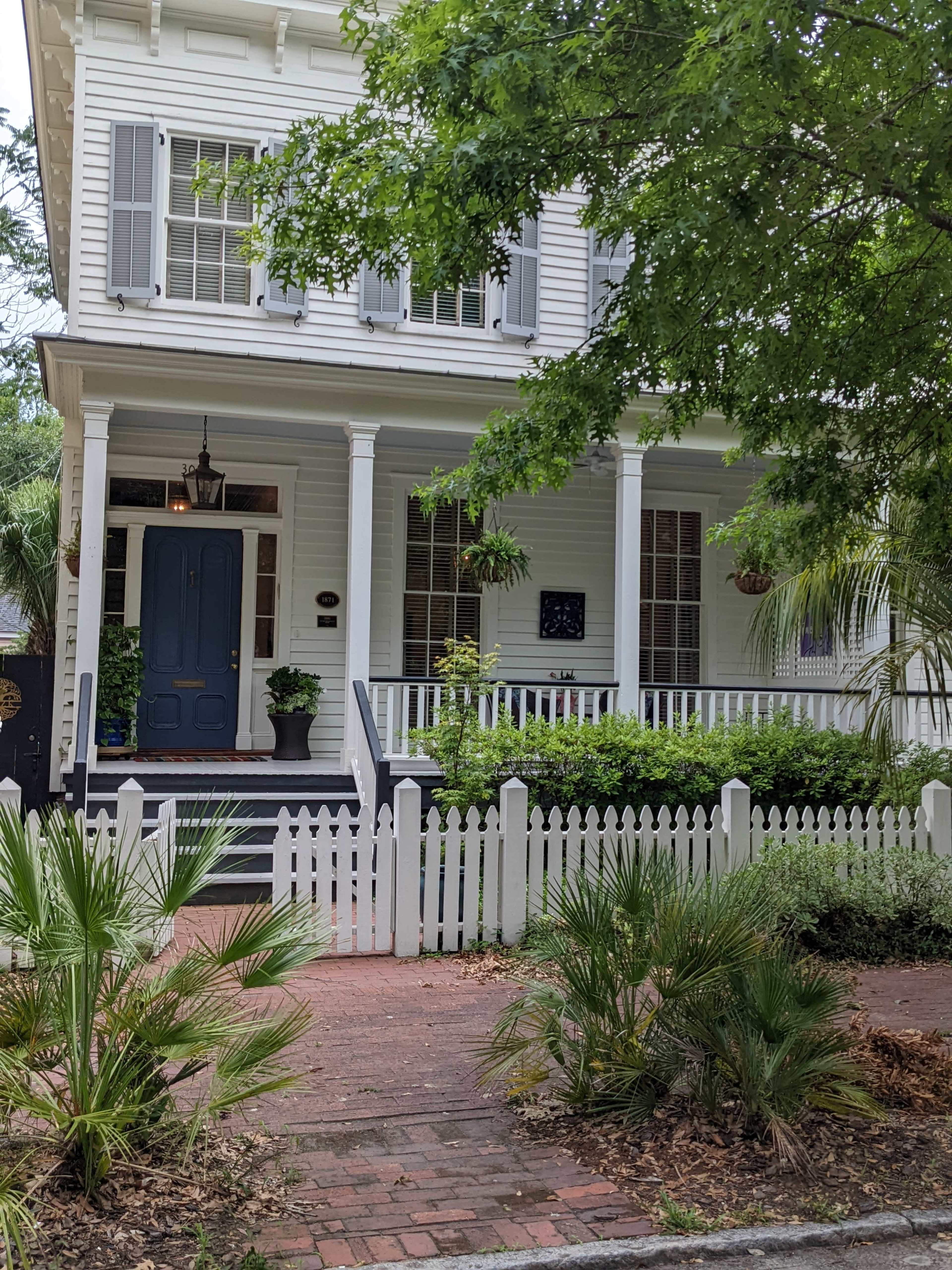 A white two-story house with a blue door, surrounded by greenery and a white picket fence.