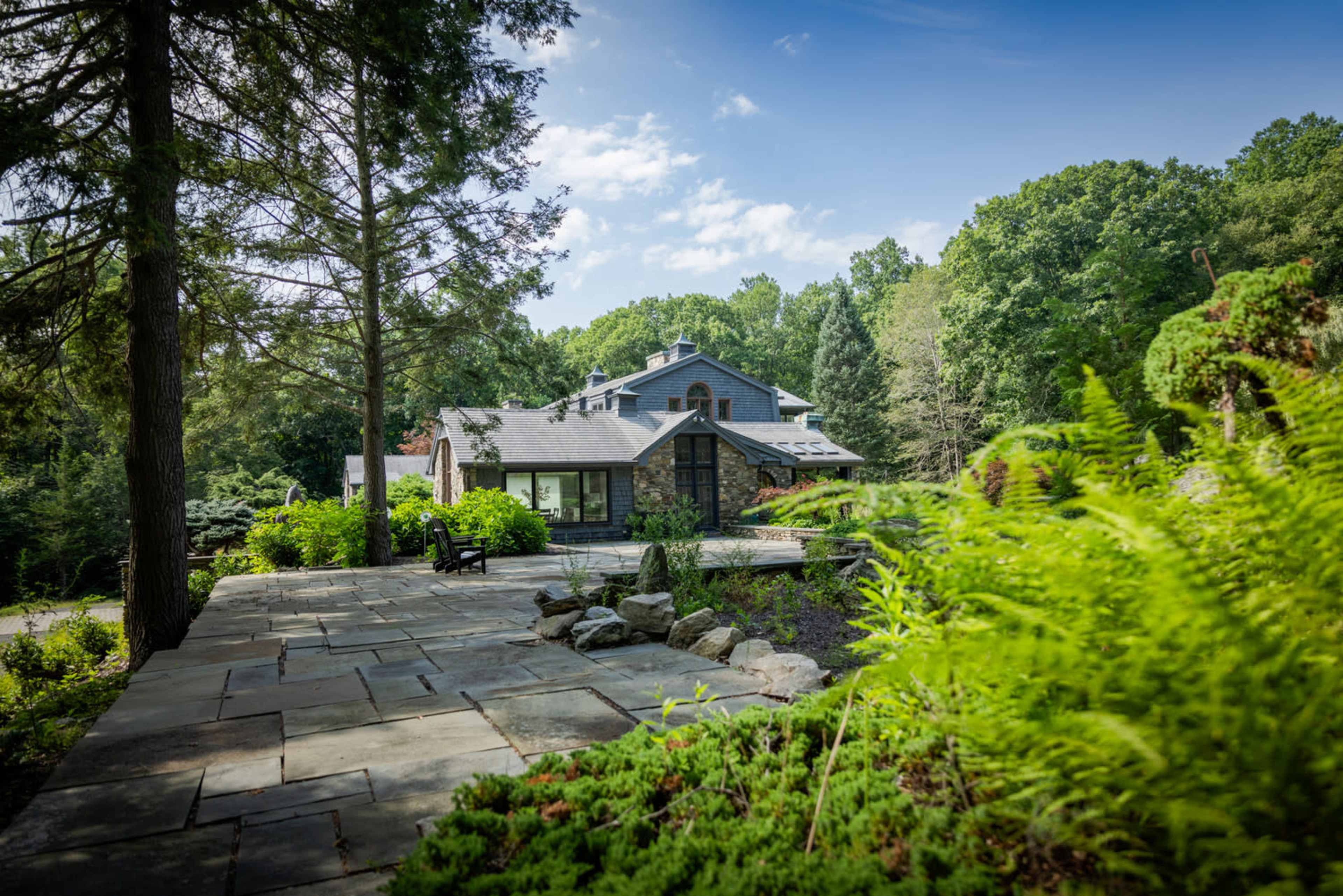 A modern stone house is surrounded by a lush green landscape and trees, with a stone pathway leading to the entrance.