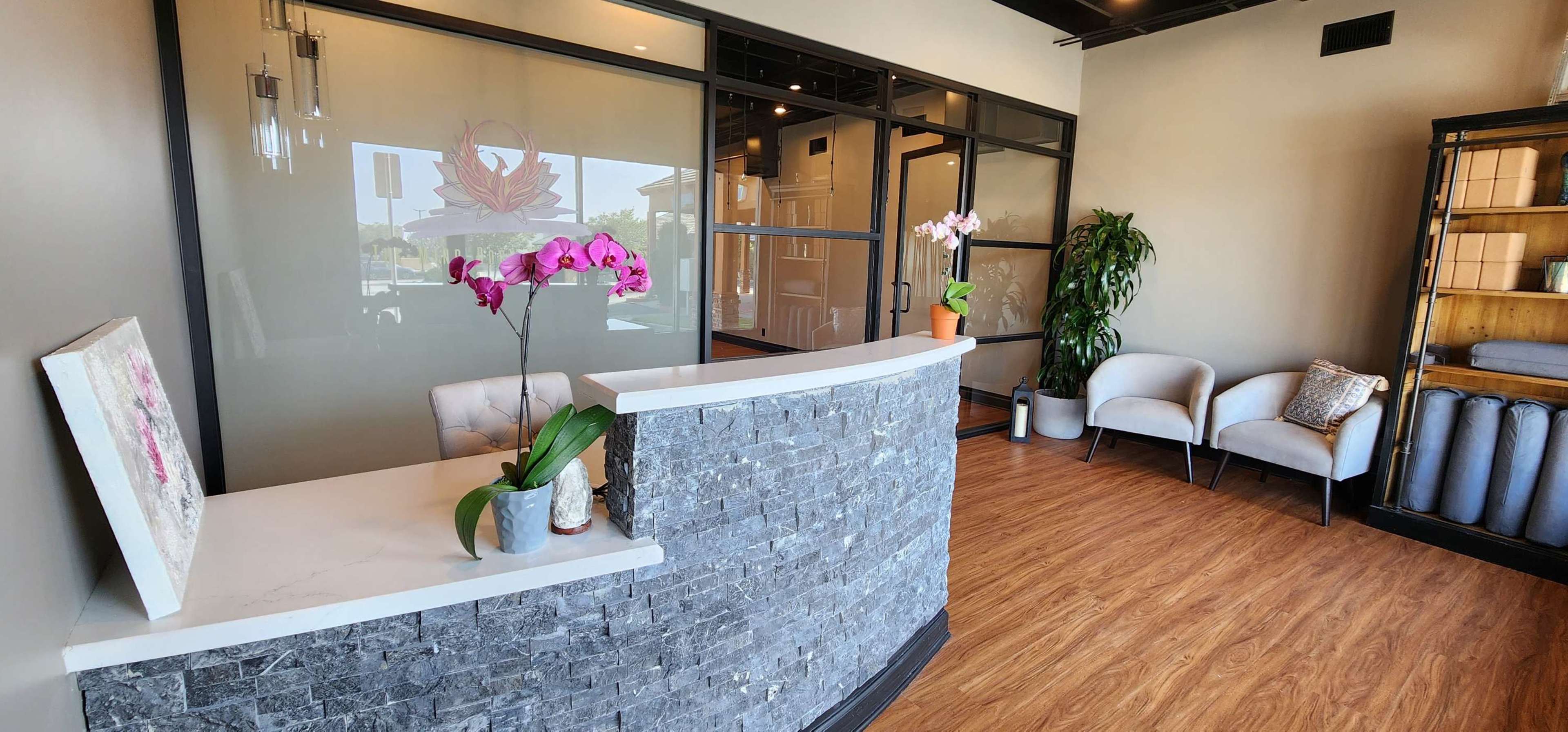 A modern reception area with a stone counter, potted orchids, and comfortable seating, surrounded by plants and shelves with rolled towels.