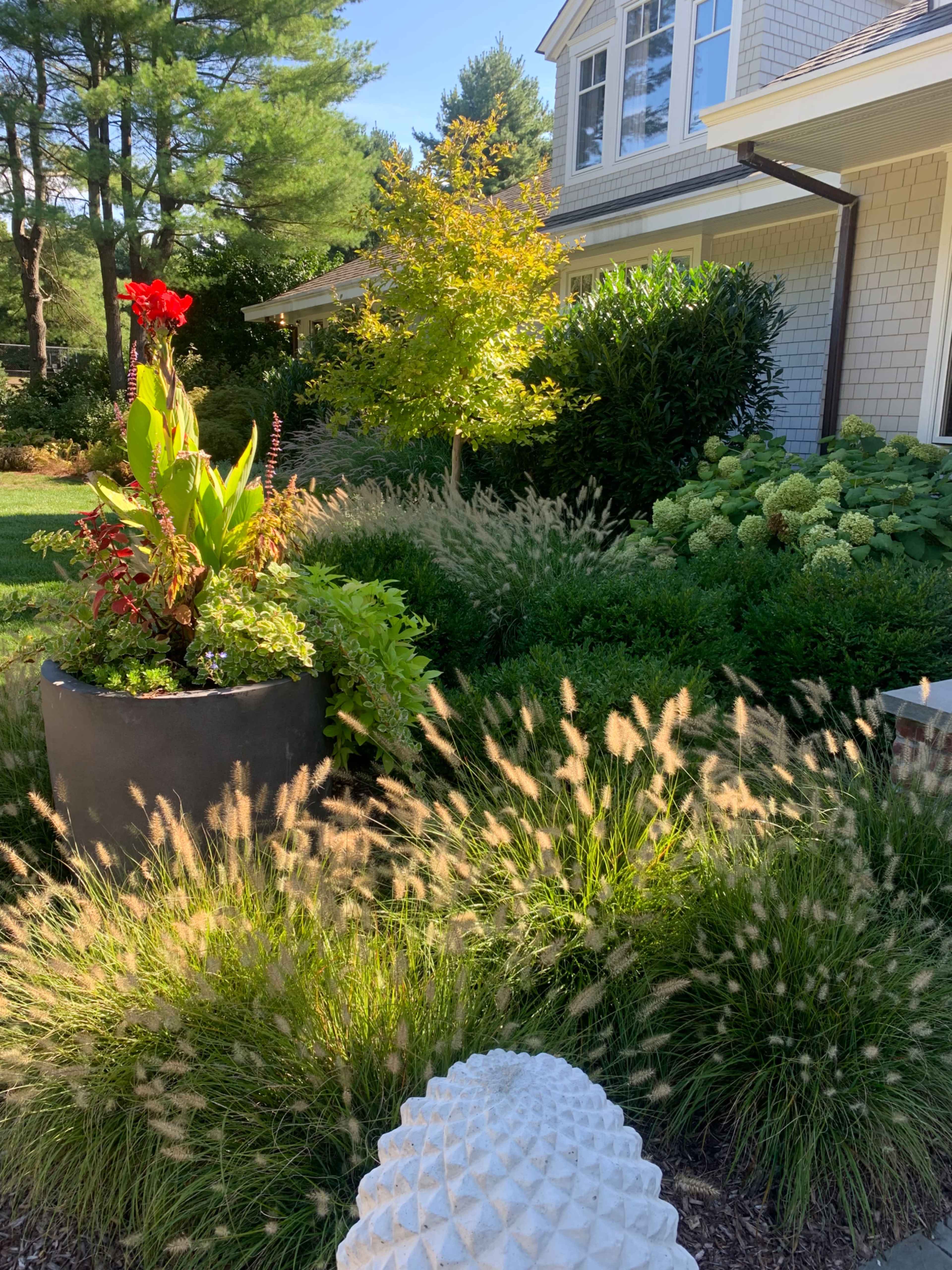 A large black planter filled with vibrant plants sits in a garden featuring various greenery and textured grasses, alongside a cream-colored decorative element.