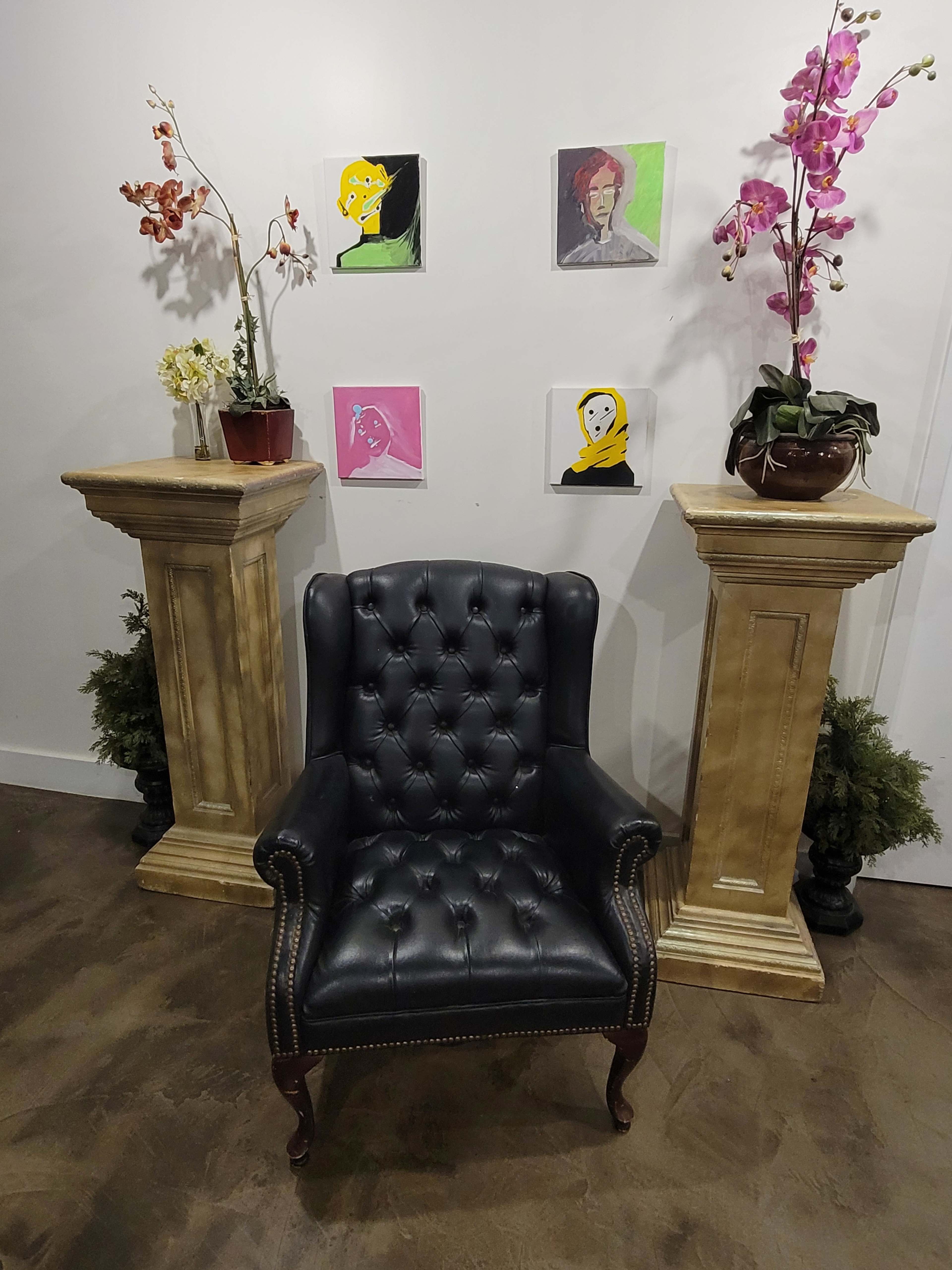 The image shows a black tufted armchair placed between two tall, decorative pedestals, with colorful art pieces hanging on the wall above and potted plants on the pedestals.
