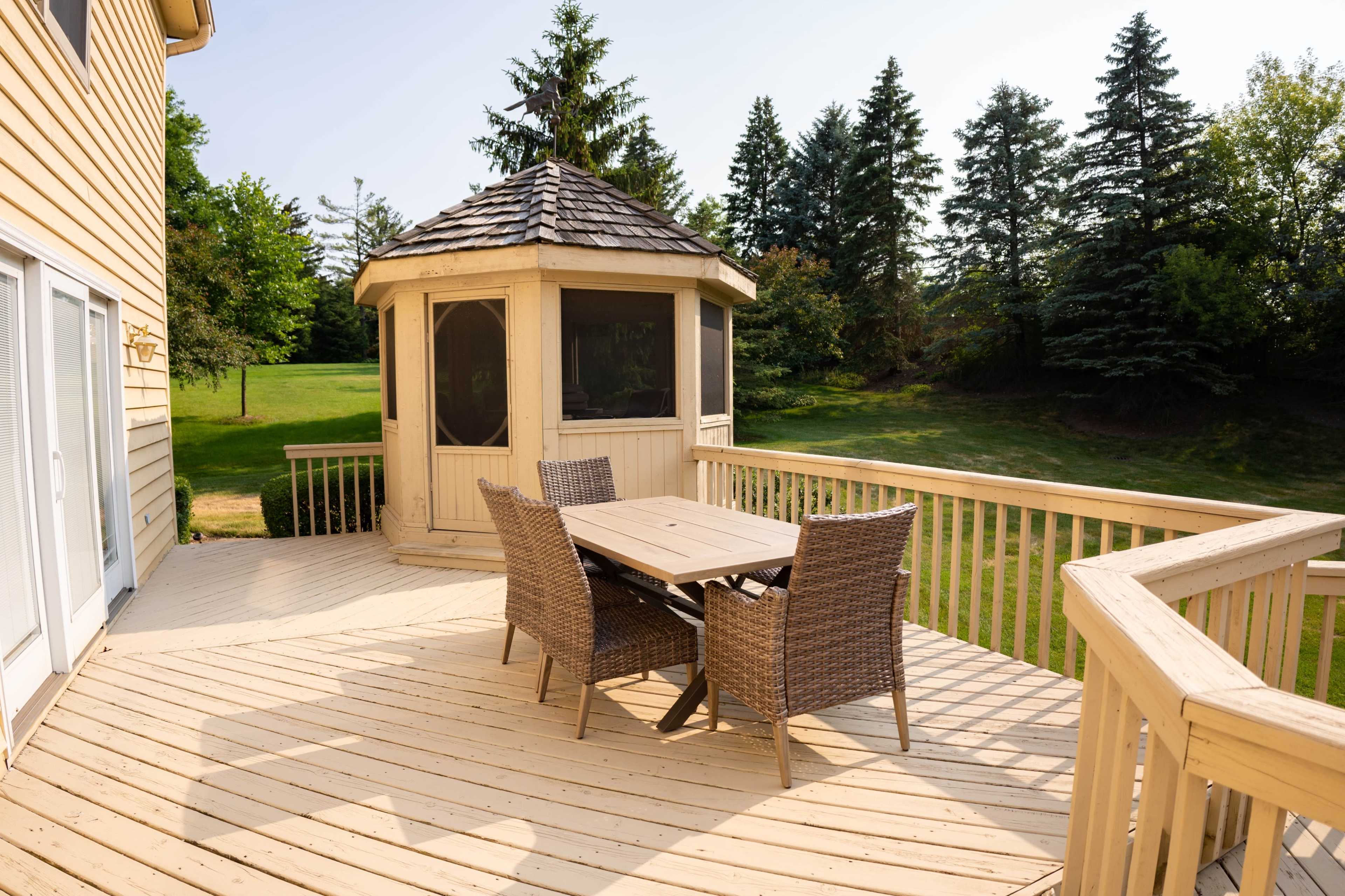 A wooden deck with a table and chairs overlooks a gazebo and a grassy area surrounded by trees.