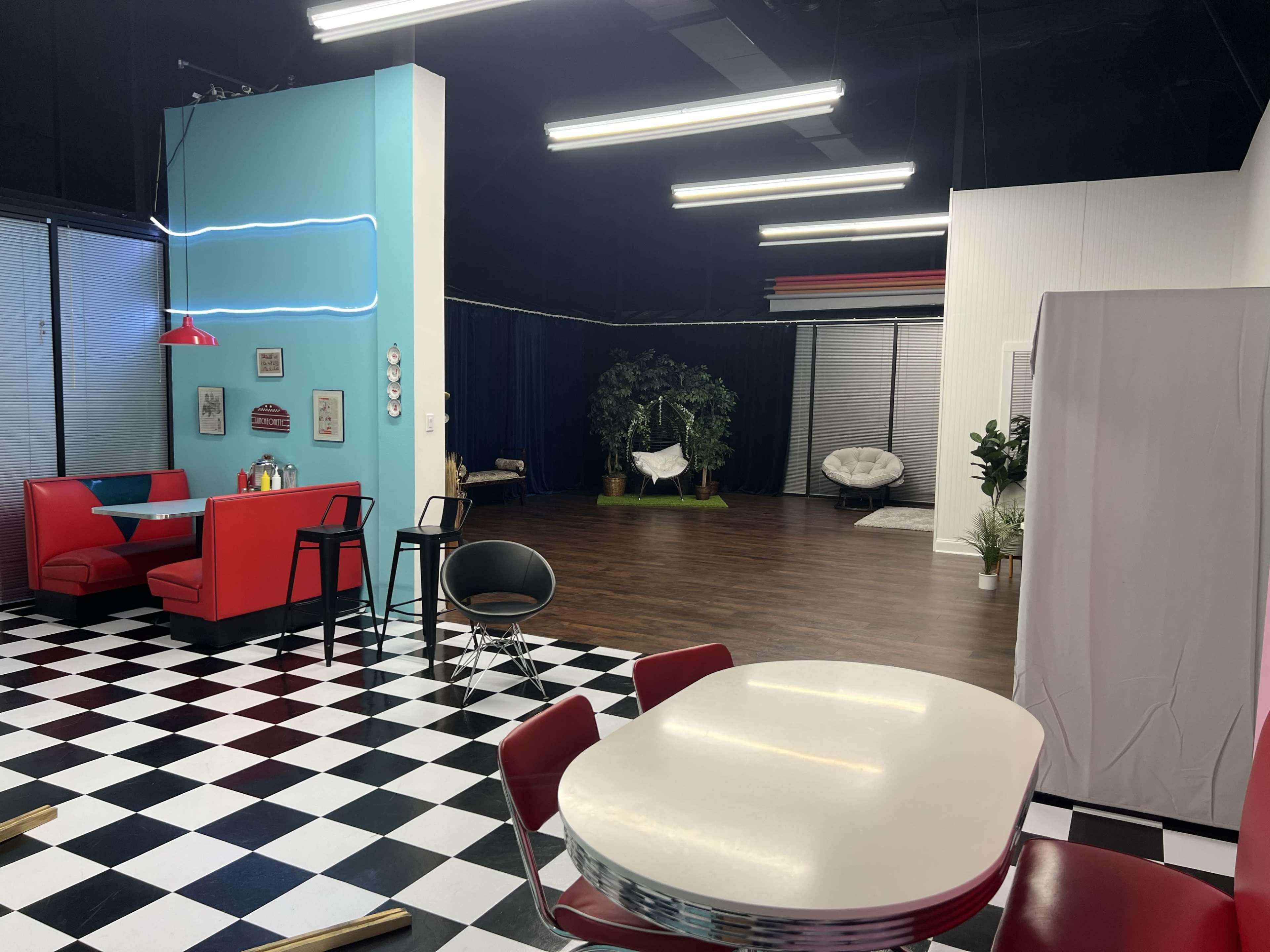 The image shows a colorful, modern interior featuring a diner-like seating area with red booths, a checkered floor, and a separate space in the background with plants and a hanging chair.