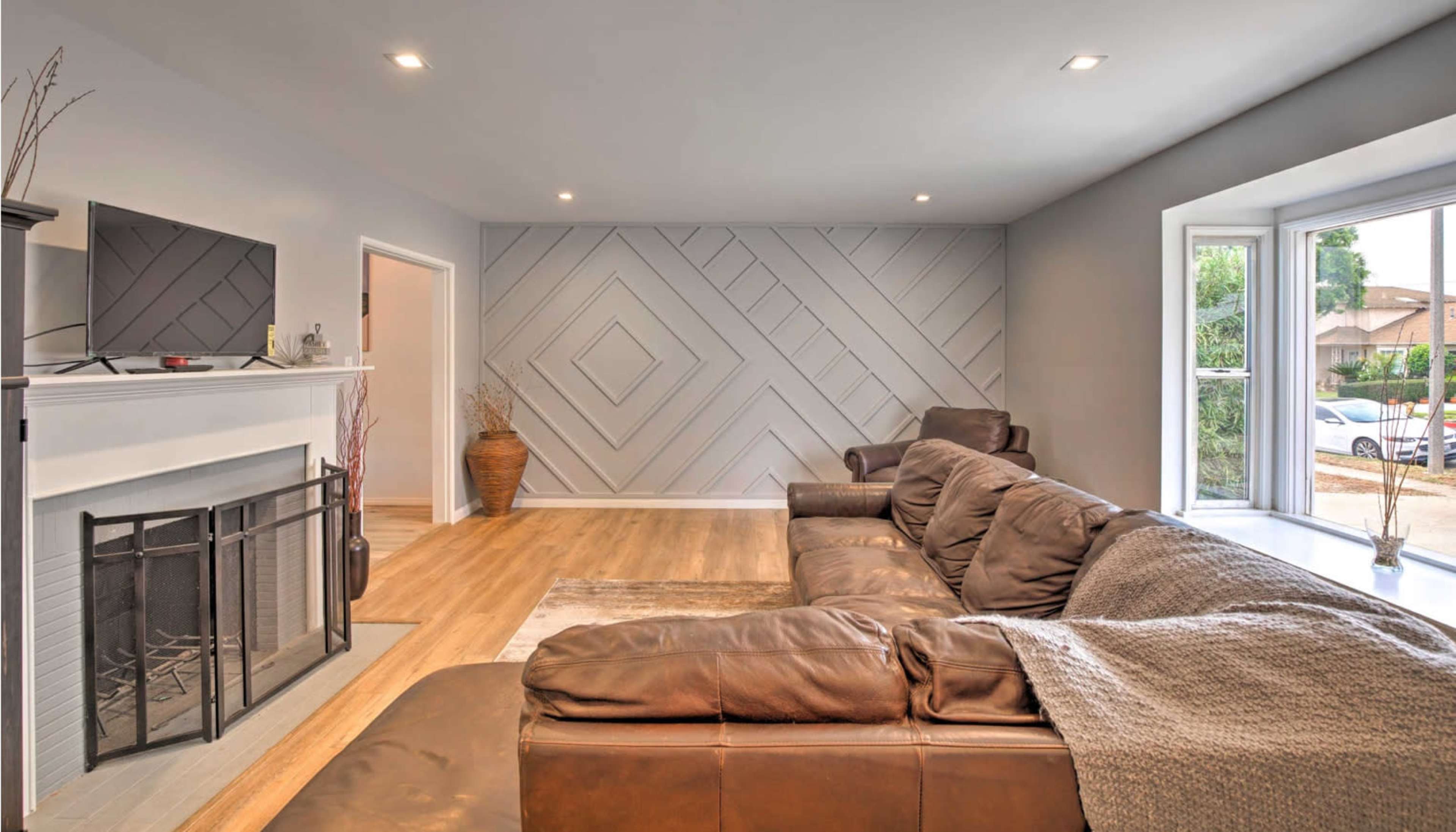A spacious living room features a brown leather sofa, a television mounted on the wall, and a large window beside a fireplace, with geometric patterns on the wall.