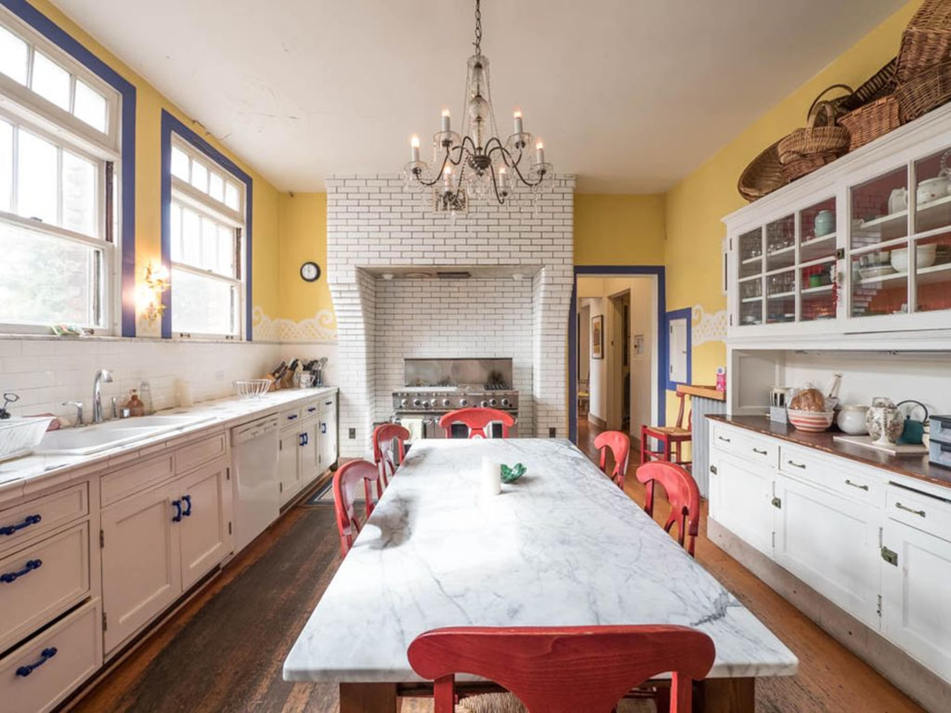 A spacious kitchen features a marble dining table surrounded by red chairs, with a white tiled fireplace and cabinets along the walls.