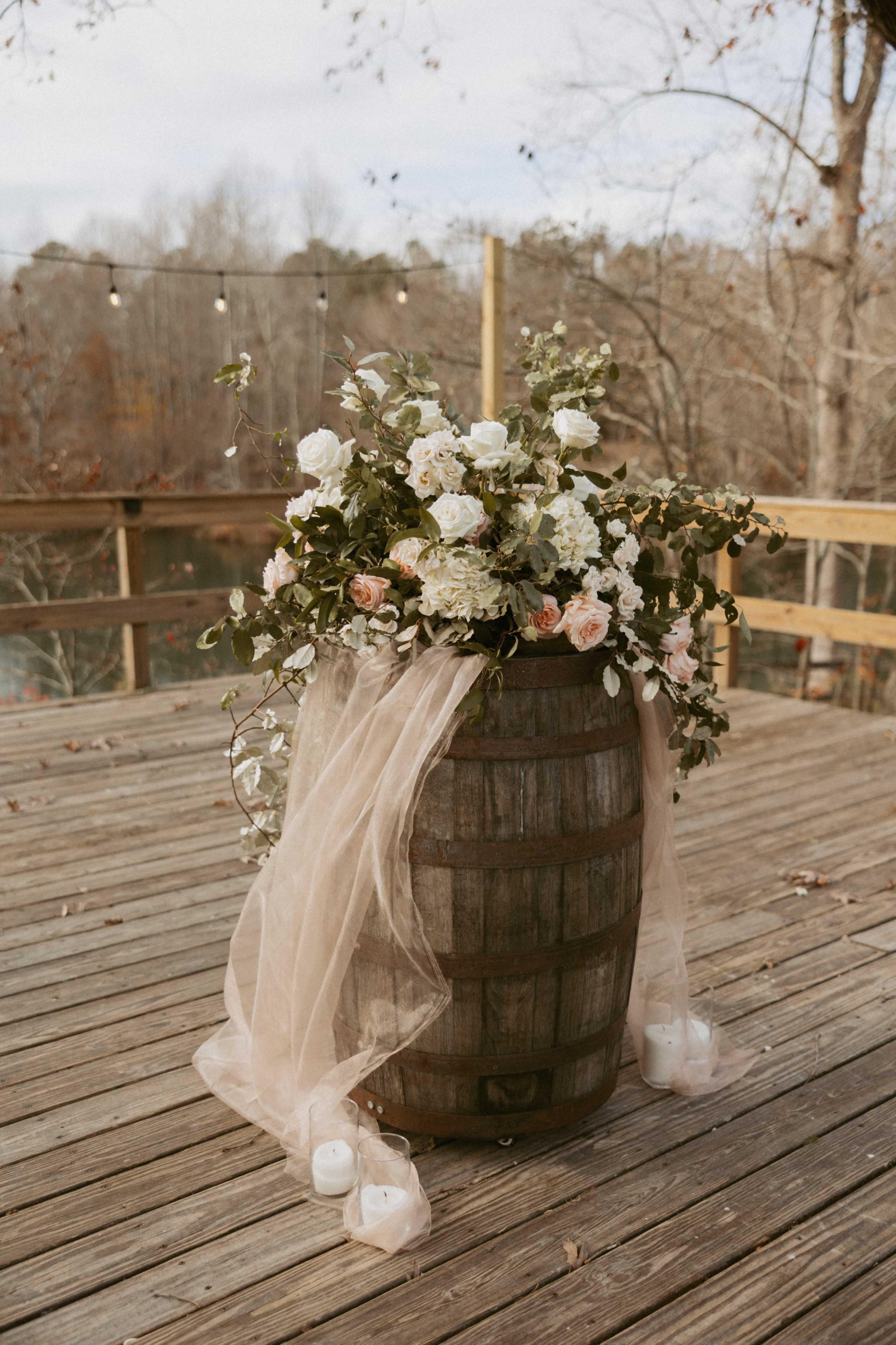 A wooden barrel is decorated with a floral arrangement and draped fabric, placed on a wooden deck near a serene outdoor setting.